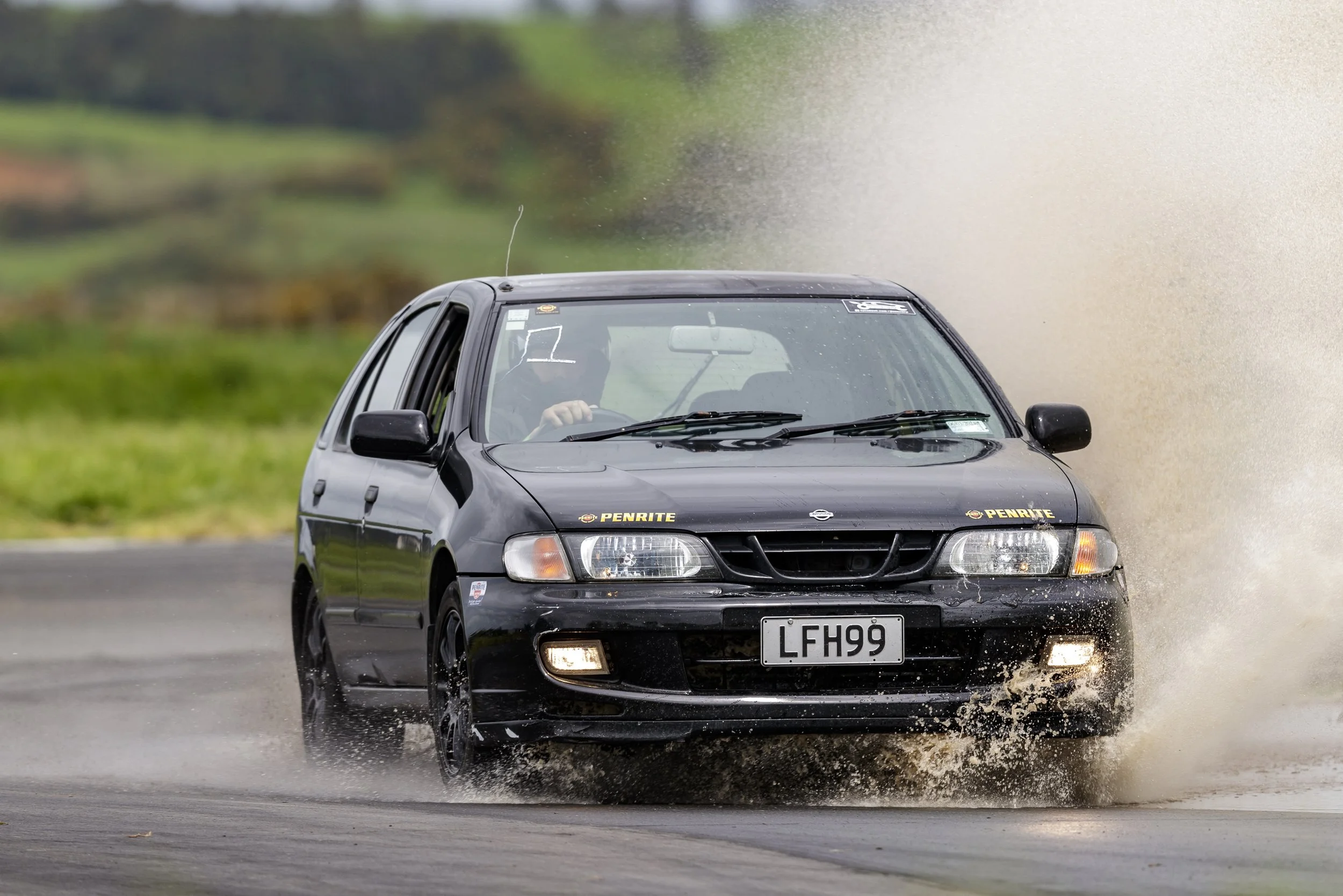 A black Nissan car driving through a shallow water puddle, creating splashes. The car has a 'Penrite' sticker on the front bumper, and the windshield has some writing on it. The background is green and blurred.