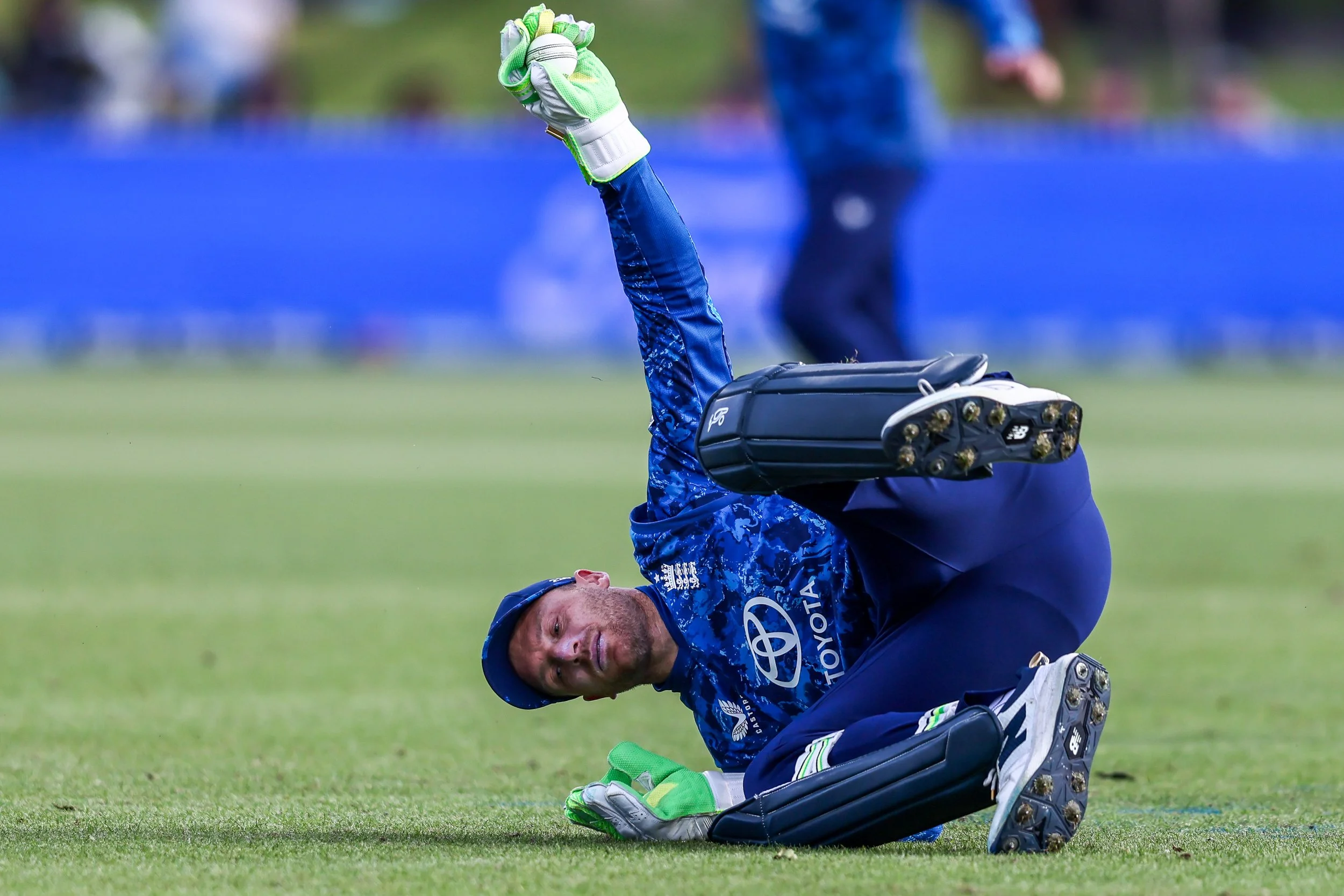 A cricket player wearing blue uniform and gloves is falling on the grass field after a game or play, with a captured position on the ground.