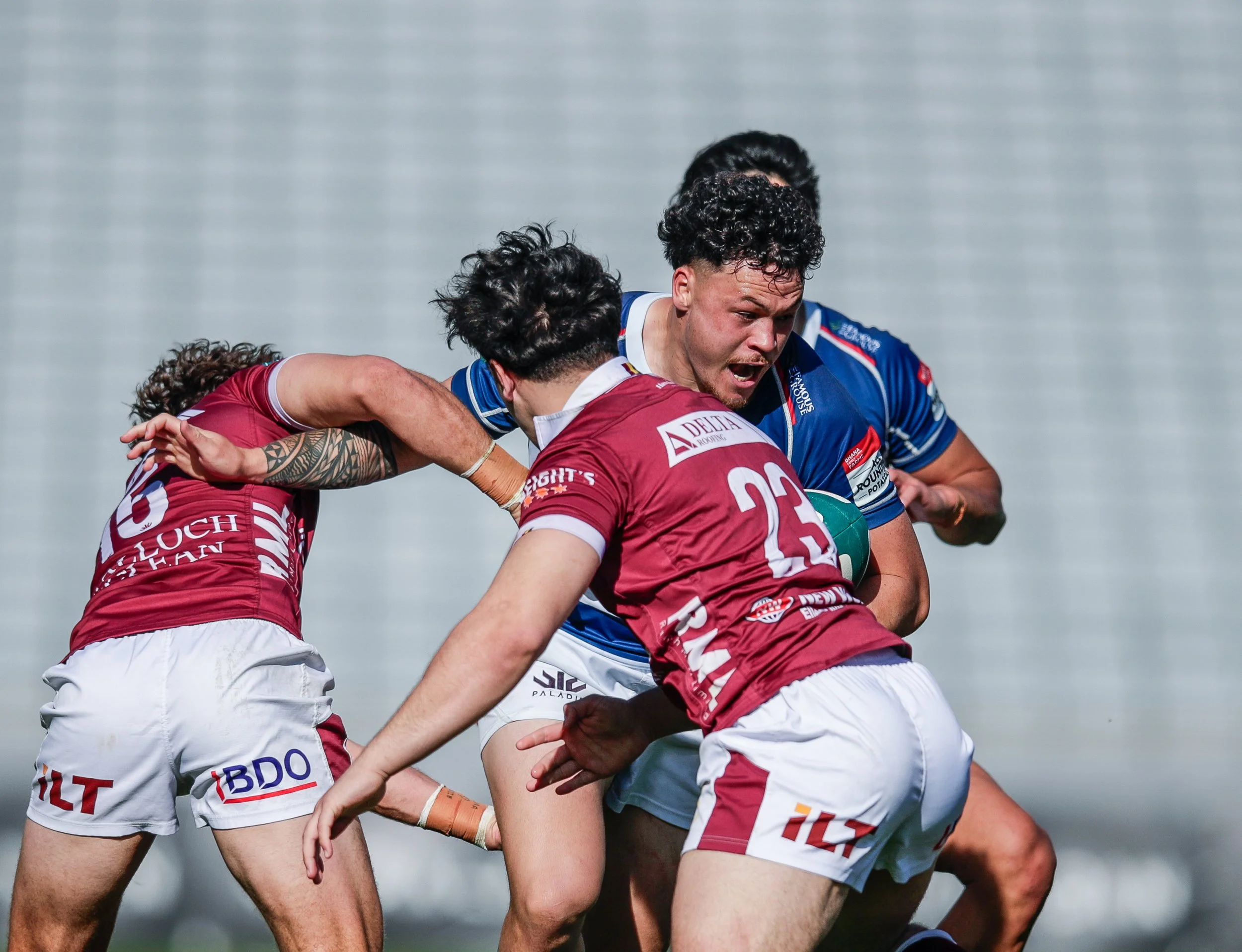Rugby players in red and blue uniforms engaged in a tackle during a rugby match on a grassy field.