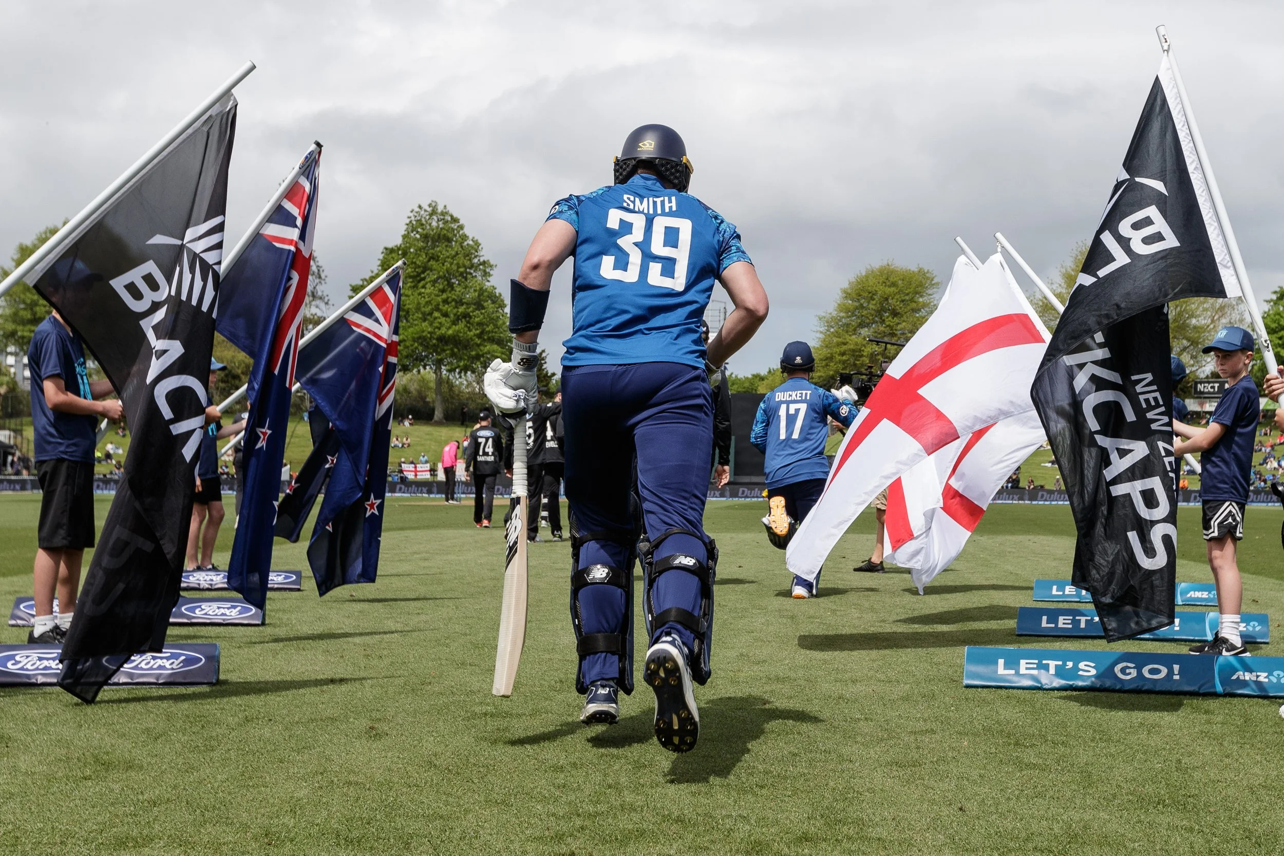 An athlete in a blue uniform with the name 'Smith' and number '39' walking on a sports field, surrounded by flags and supporters, near a sign that says 'Let's Go!'.