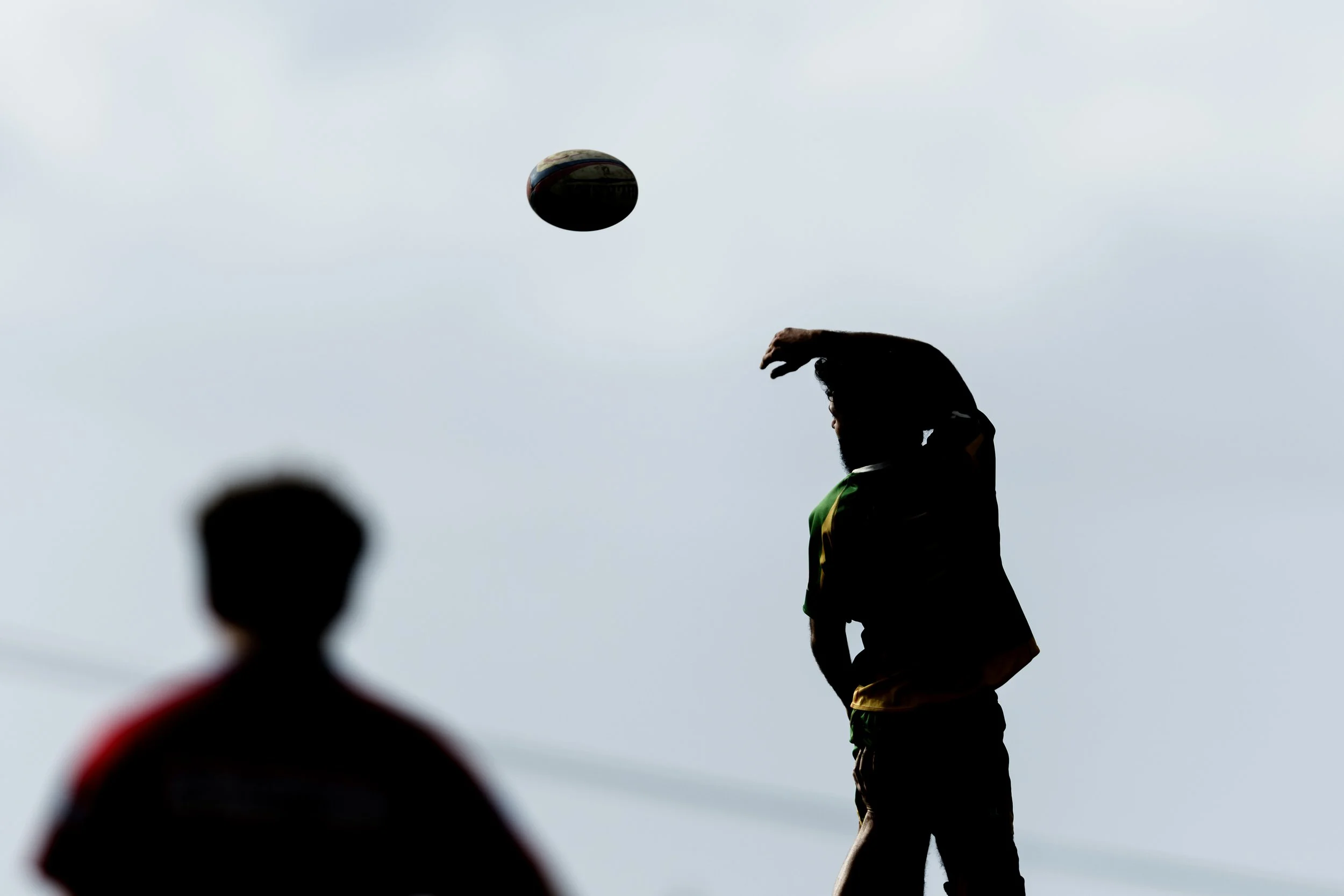 A person playing rugby, with a ball in mid-air and their hand raised as if about to hit the ball. There is a blurred figure in the foreground and overcast sky in the background.