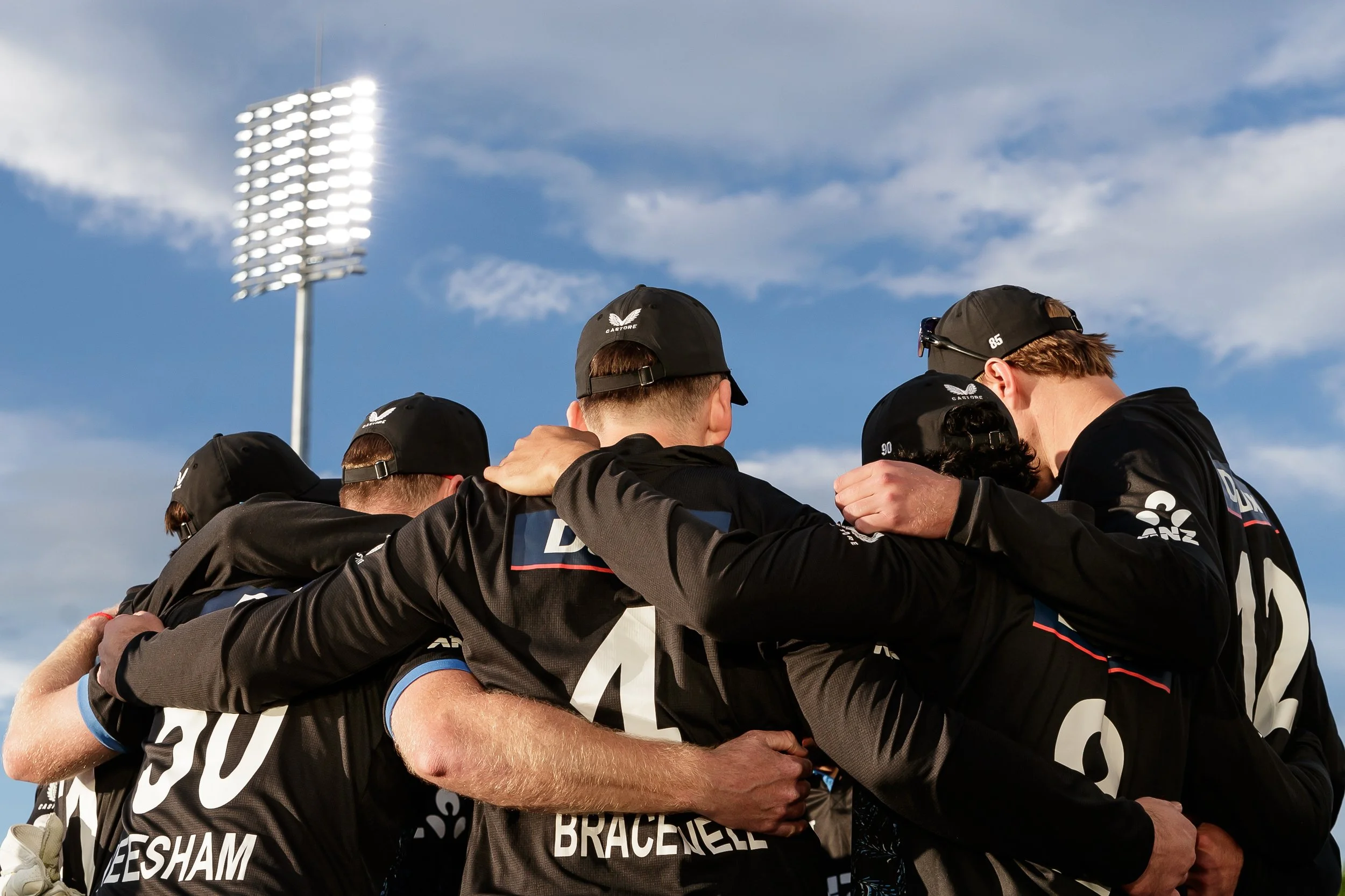 A group of athletes wearing black sports uniforms and black caps with team logos, huddled together with arms around each other on a sports field under a partly cloudy sky.