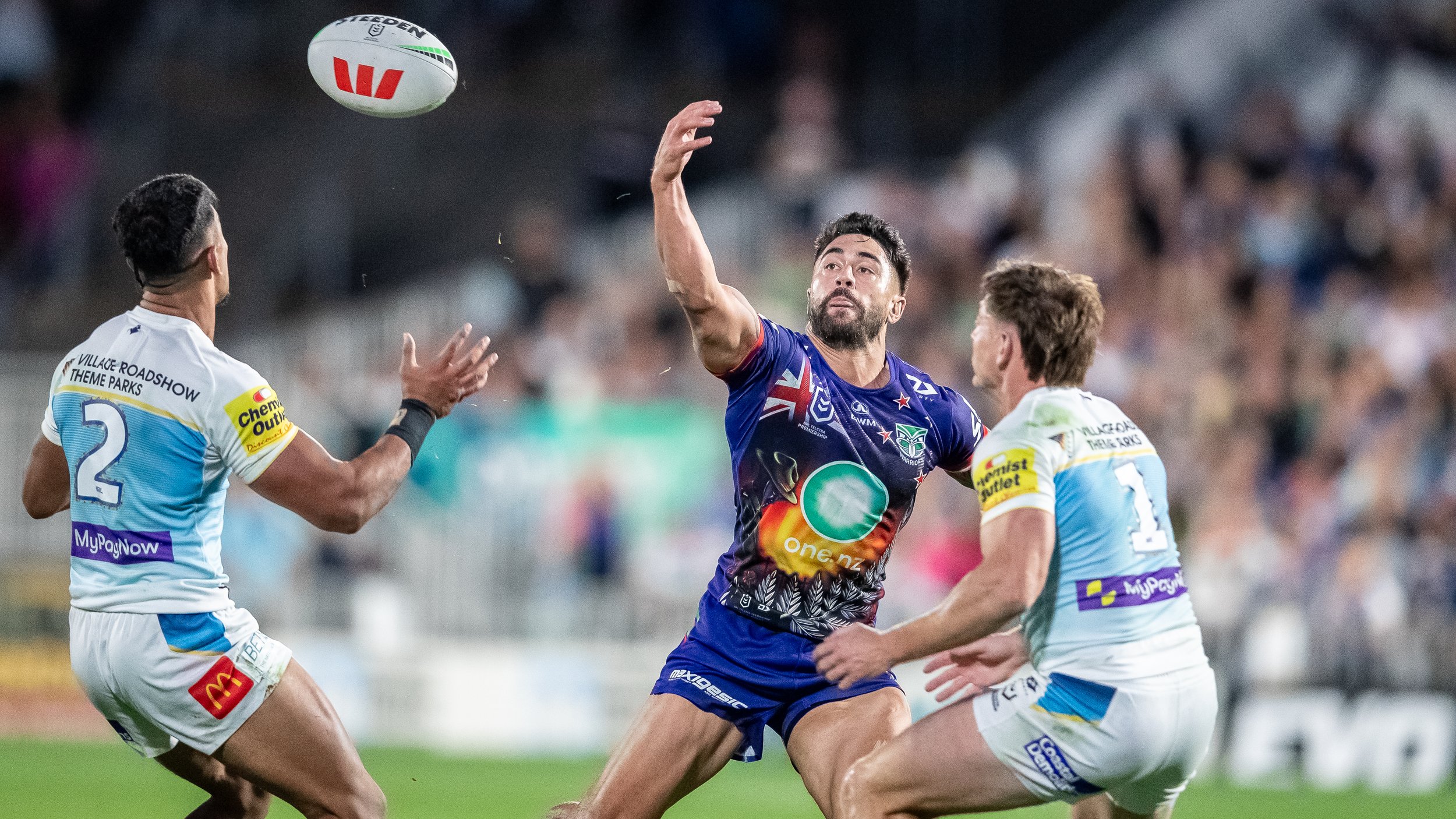 Three rugby players in action during a game. The player in the middle is reaching to catch the ball, while two players on either side are defending.