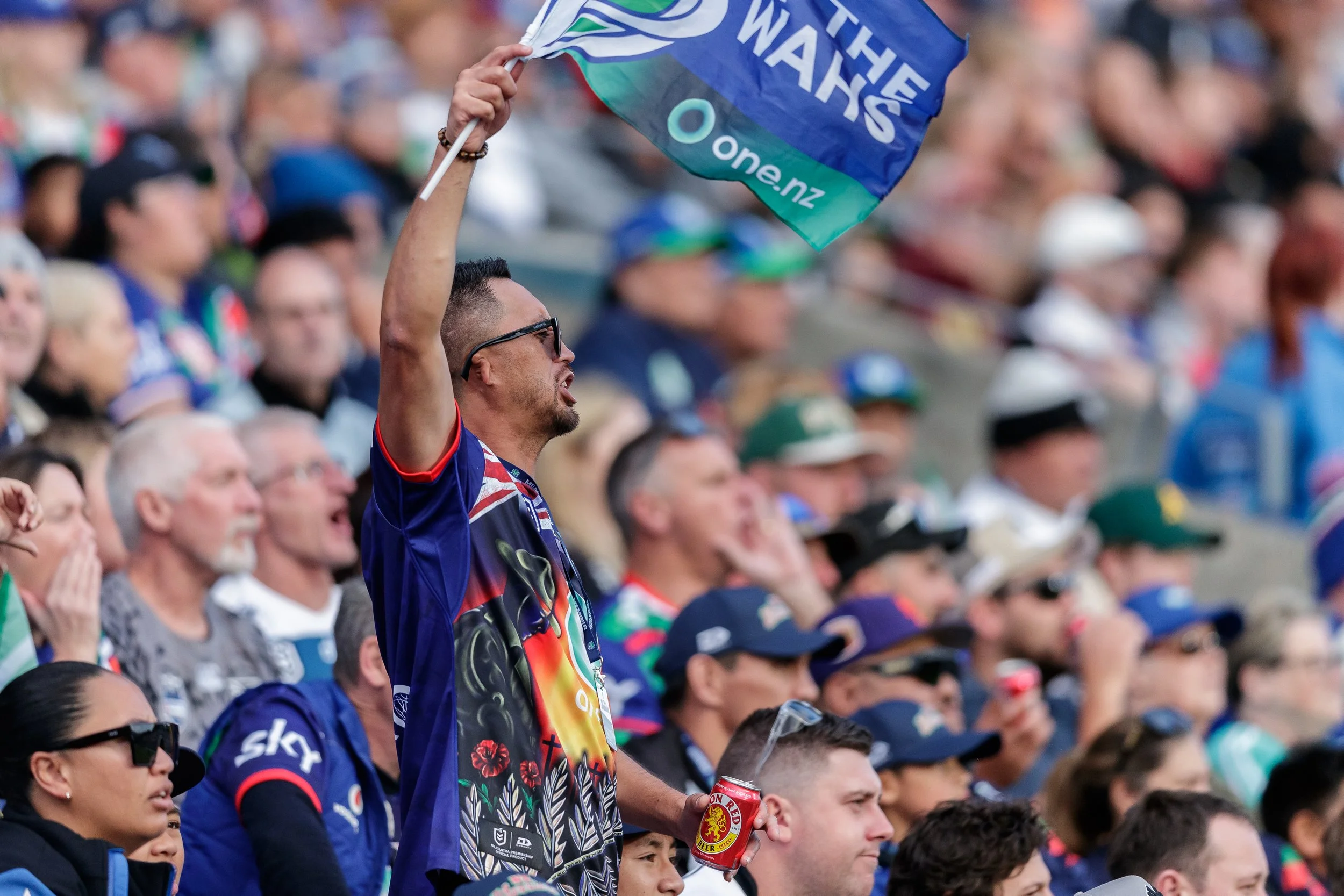 A man wearing glasses and a colorful sports jersey is standing among a crowd, holding a can of Coopers beer and waving an Australian flag at what appears to be a sports event.