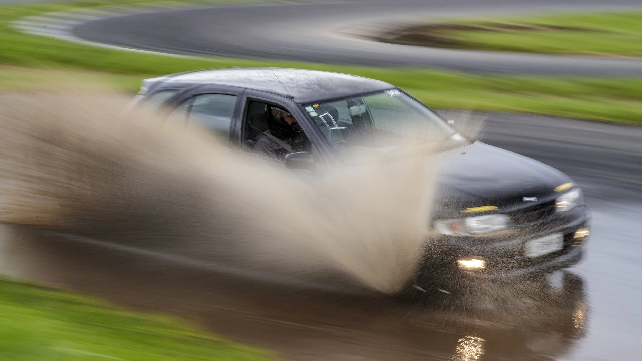 A black car racing on a wet race track, creating a splash of water as it speeds around a bend with green grass surrounding the track.