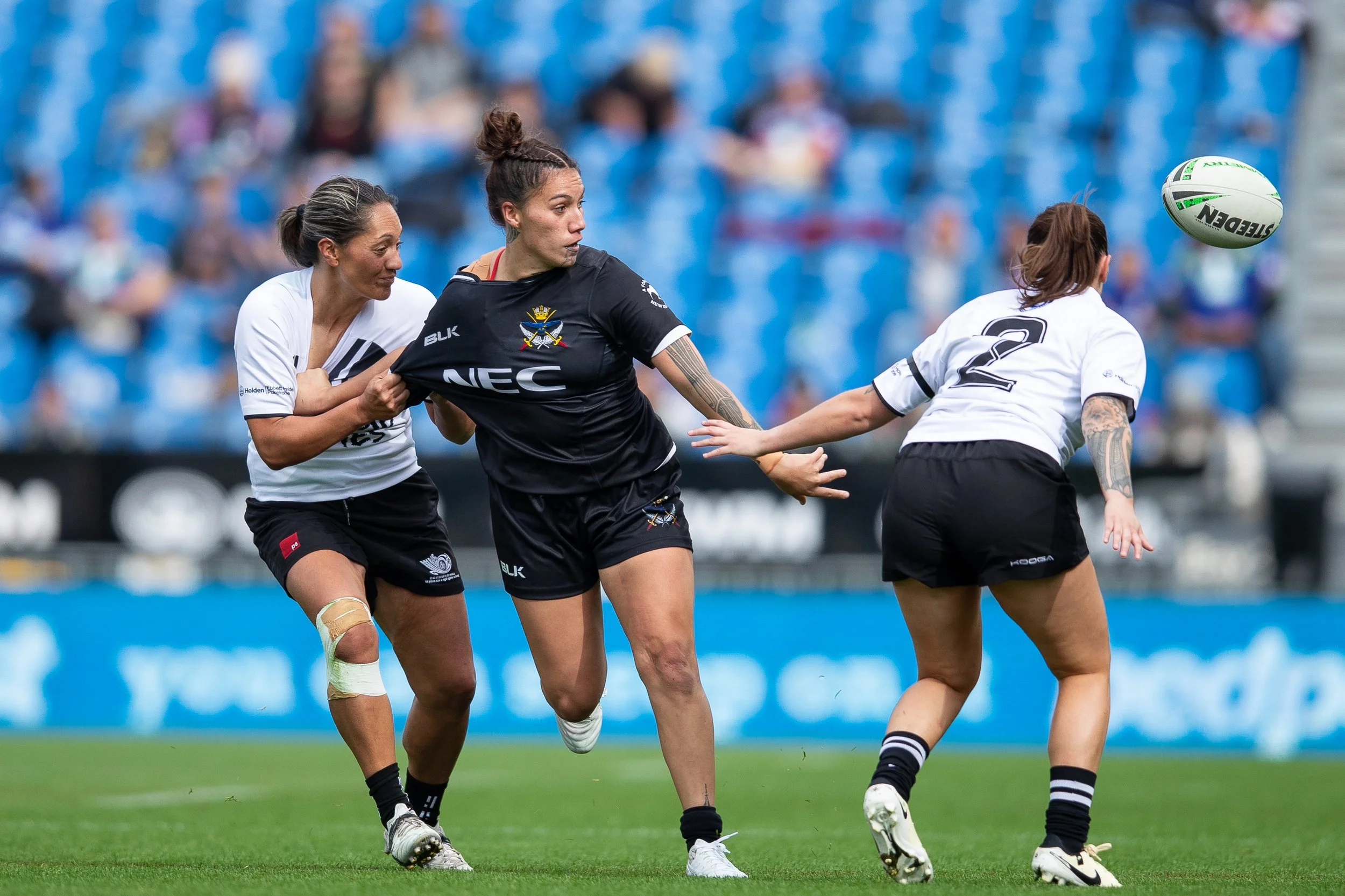 Women playing rugby on the field, with one holding the ball and others reaching out to contest for it, in a stadium with blue seats.