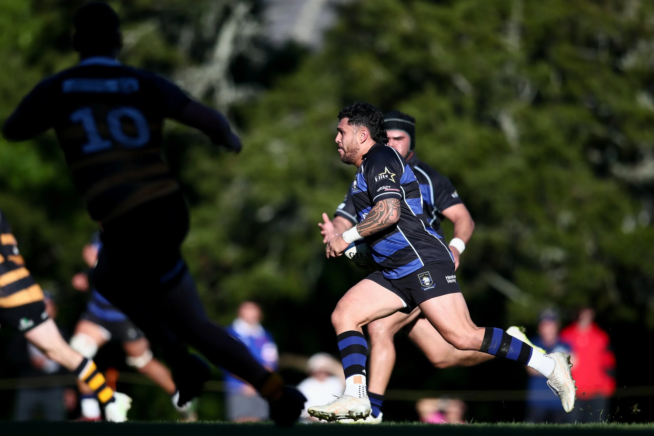 Rugby players in black and blue uniforms playing on a grassy field with trees in the background, with a focus on a player with a tattoo on his arm.