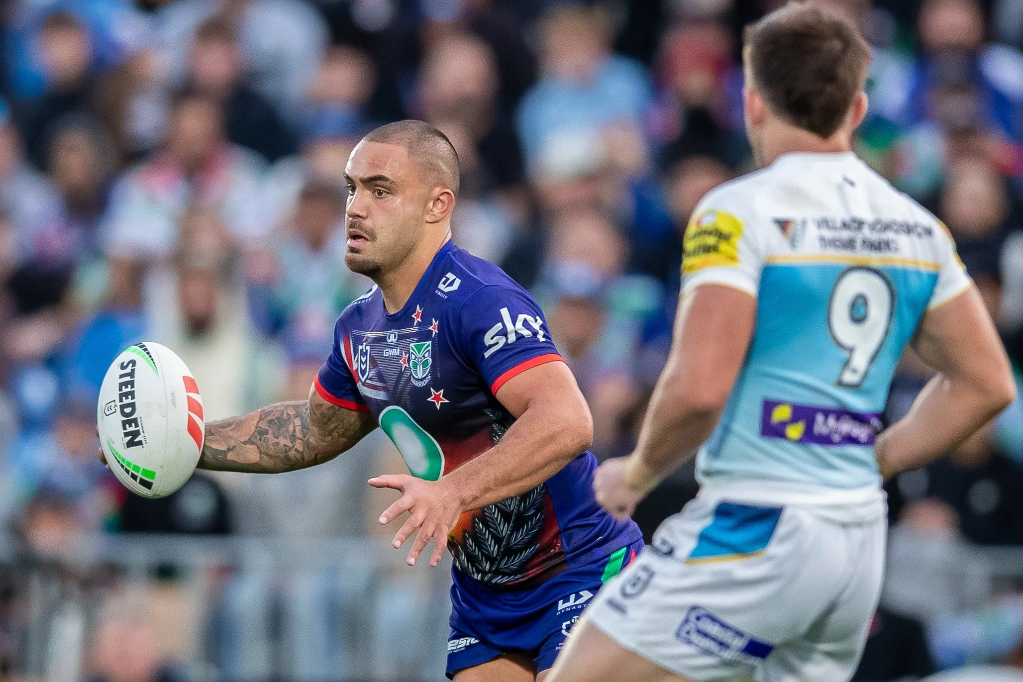 Two rugby players in action during a game, one holding the ball and the other approaching, with a blurred crowd in the background.