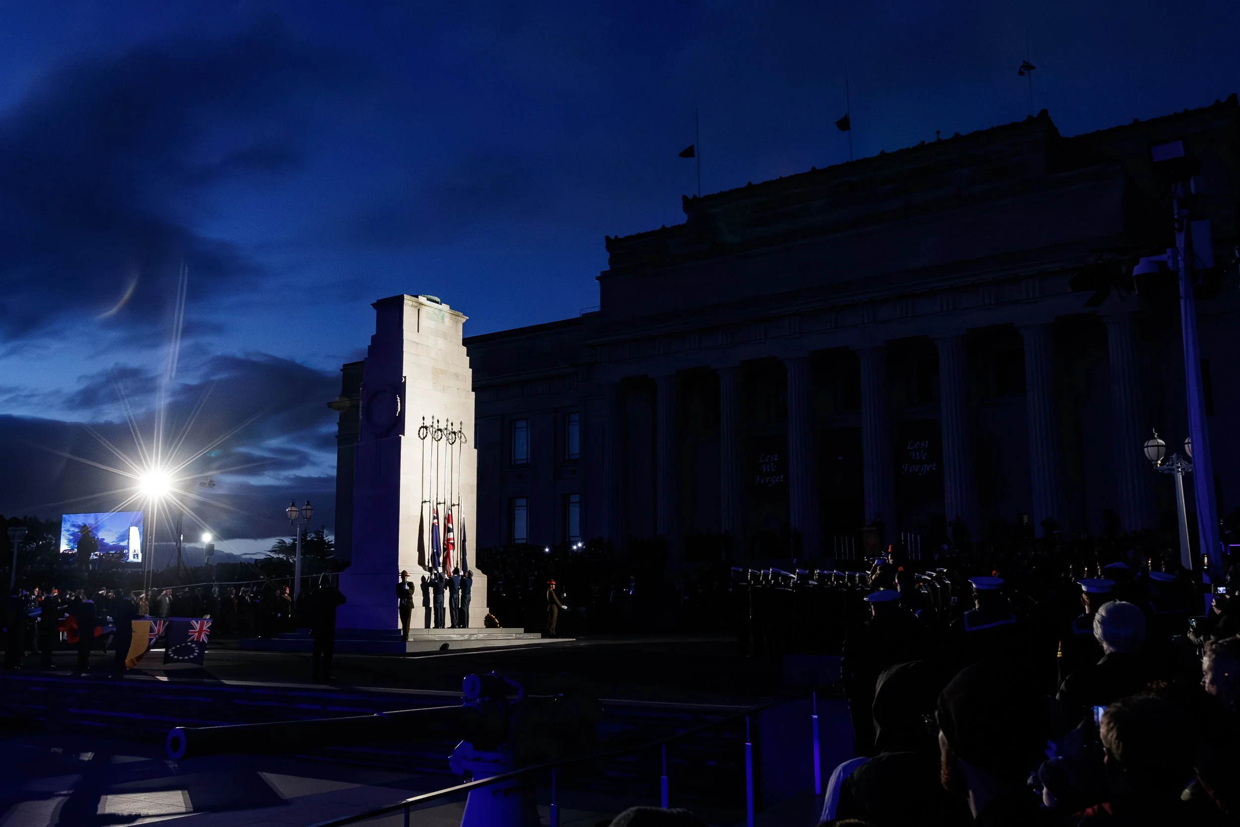 A nighttime scene at a war memorial, featuring a white stone memorial with an inscription, flags, and a crowd of people gathered for a ceremony, illuminated by bright lights against a dark blue sky.