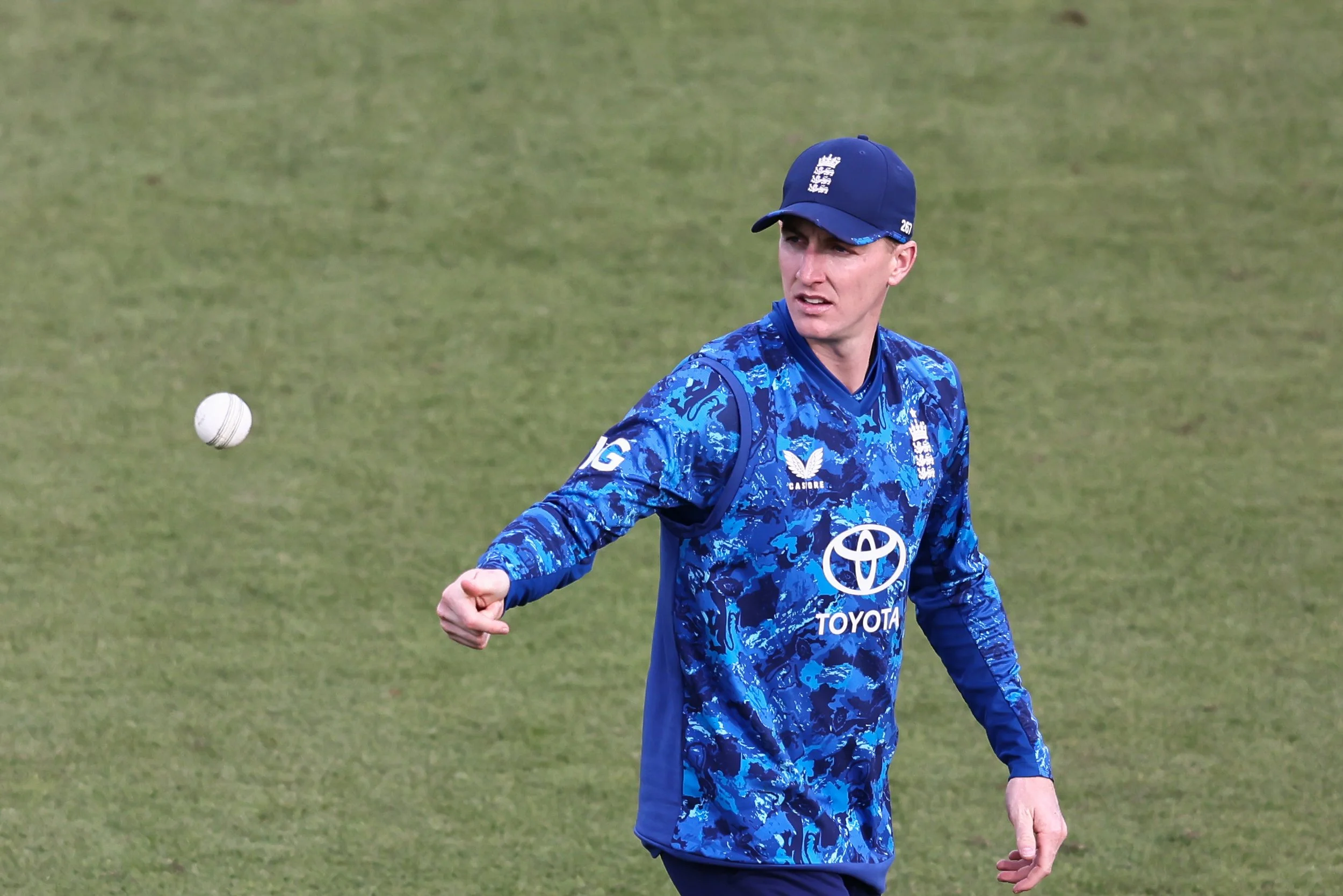 A cricket player wearing a blue uniform is on a grassy field. The player is looking to his left with a focused expression, and a cricket ball is in midair to his left.