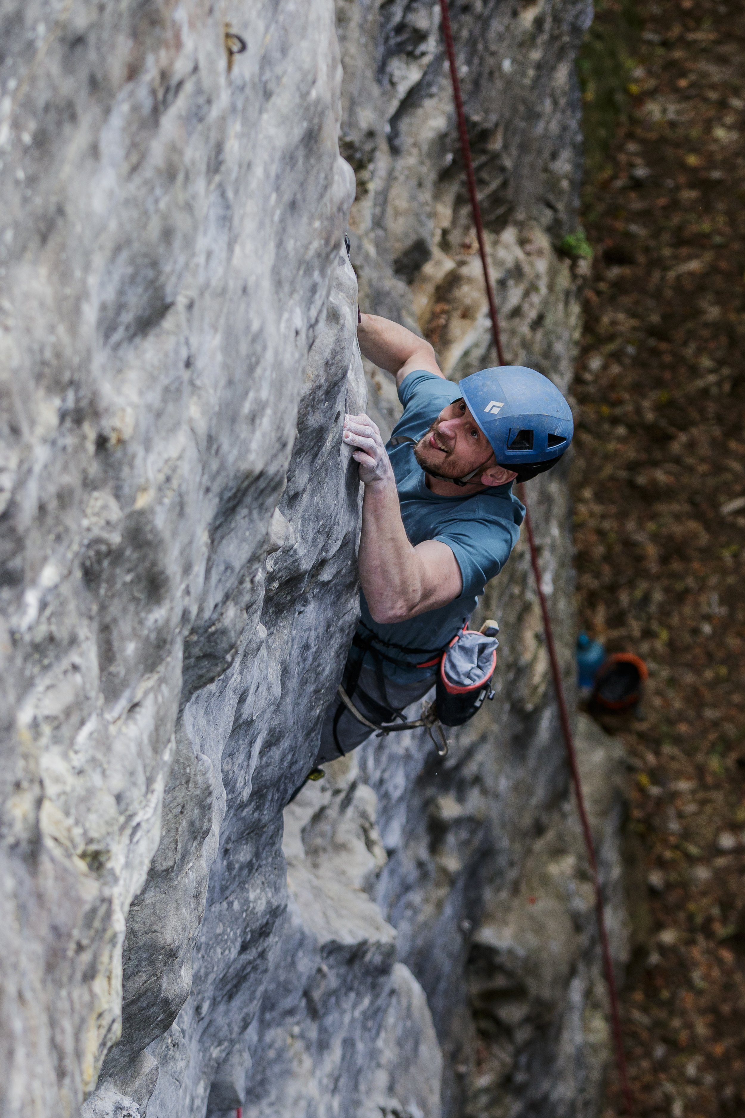 A man wearing a blue helmet and climbing gear is rock climbing on a vertical rock face outdoors. He is gripping the rock with one hand and looking up, with a red safety rope attached.