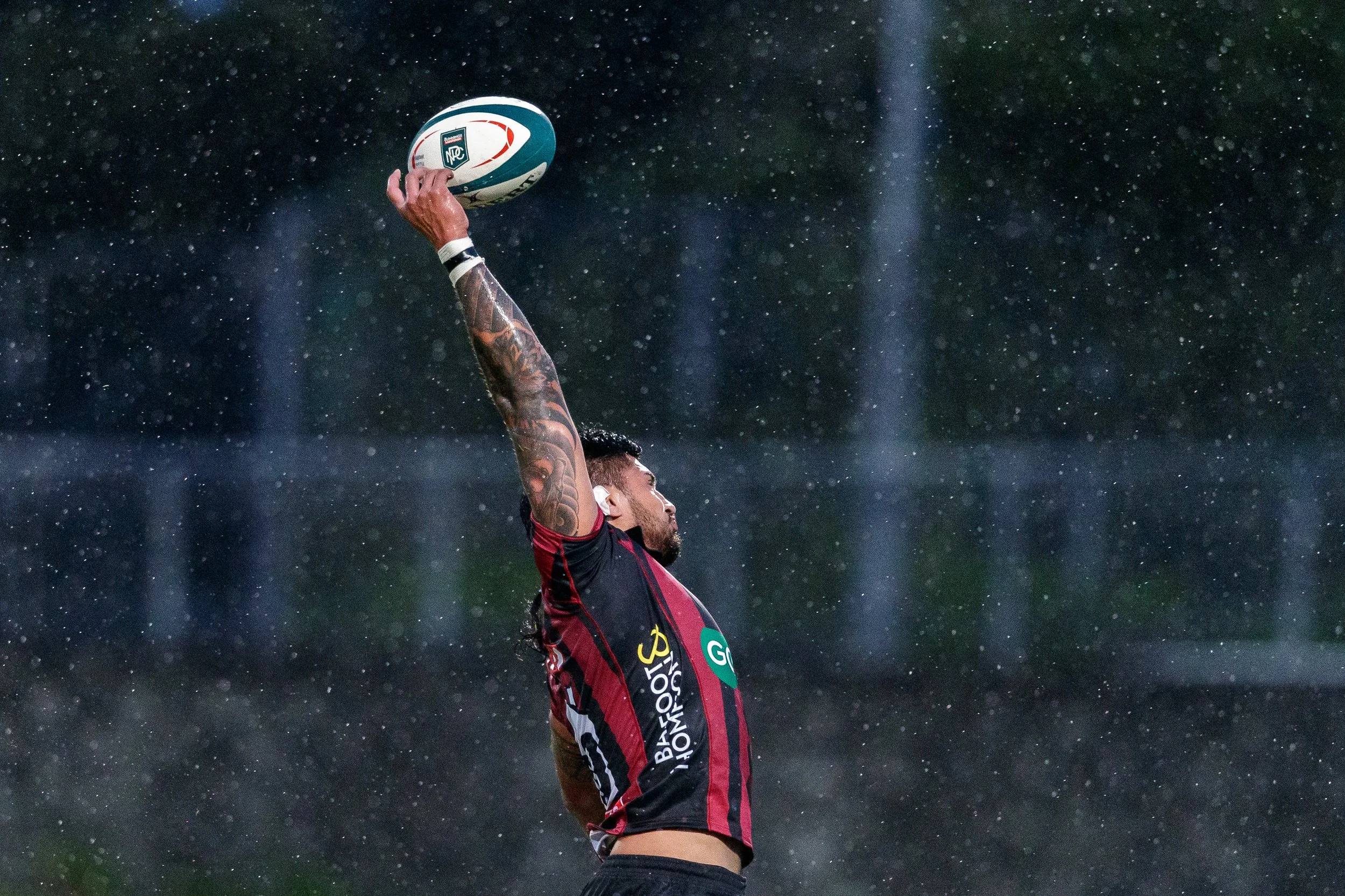 A rugby player in a black and red uniform holding a rugby ball above his head during a game in the rain.
