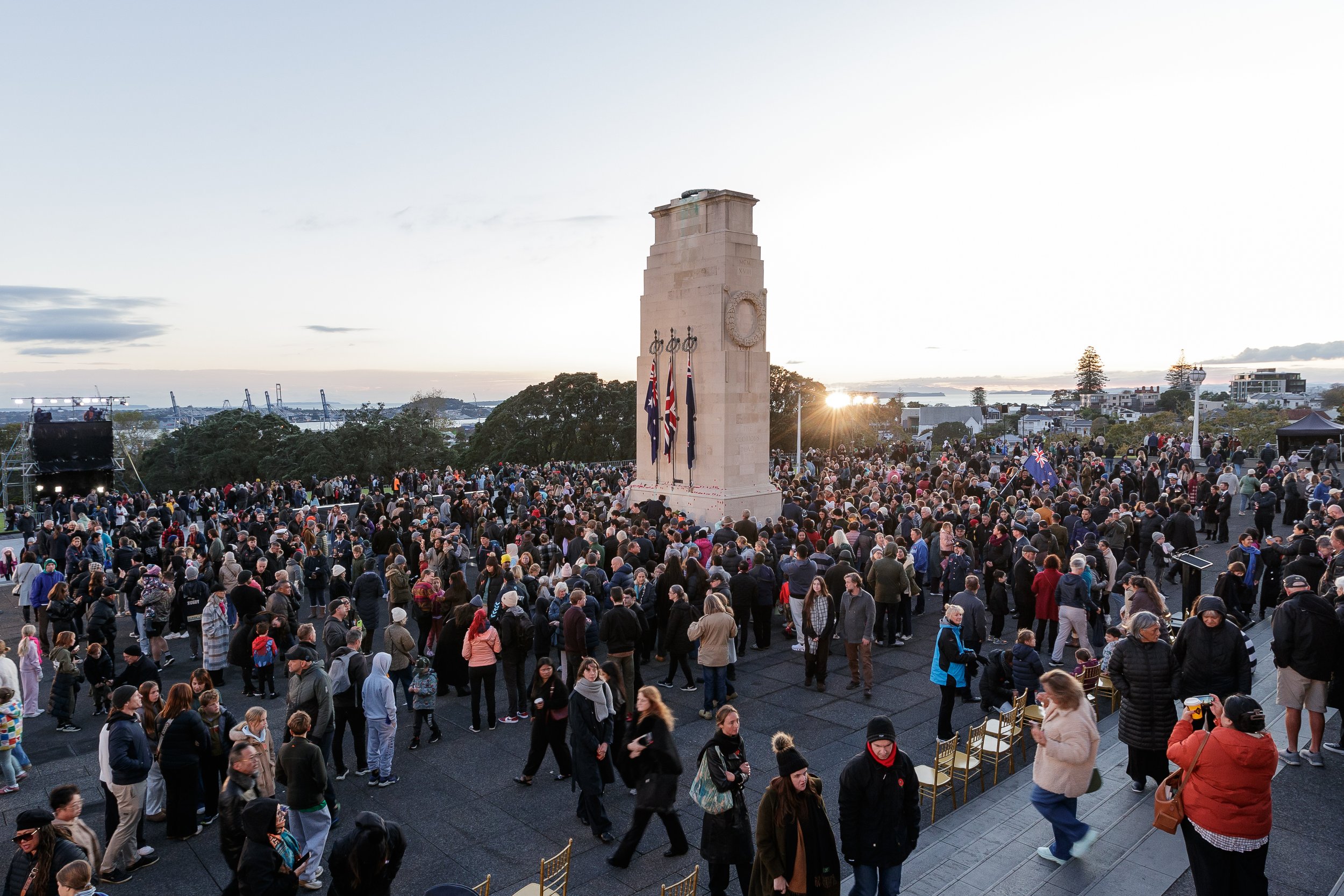 Crowd of people gathered around a monument with flags at sunset, overlooking a harbor and cityscape.
