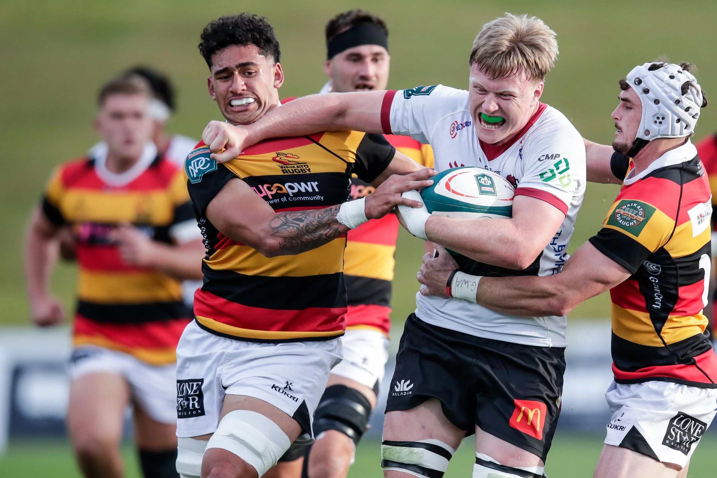 Rugby players competing for the ball during a match, with two players holding the ball while being tackled by opponents, on a grassy field.