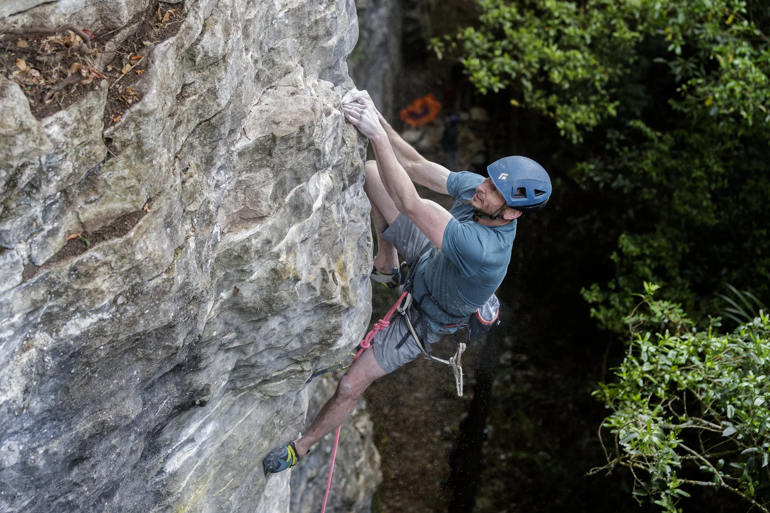 A man wearing a blue helmet and gray shorts is rock climbing on a vertical outdoor rock face surrounded by green foliage.