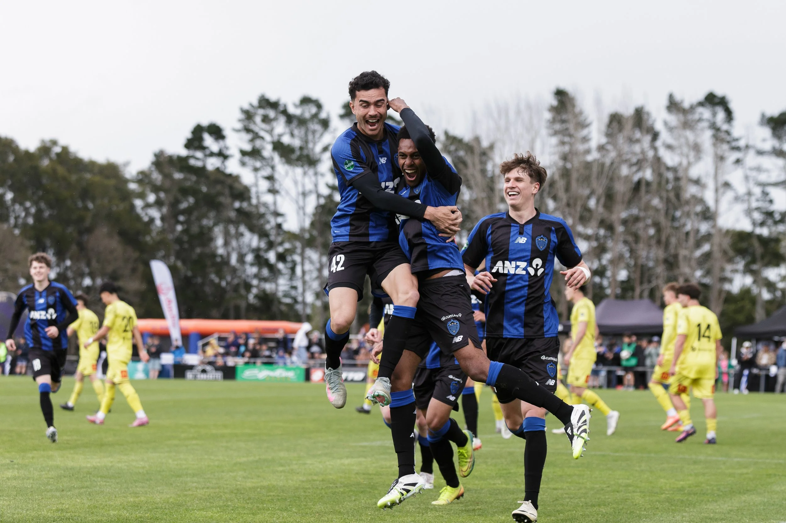 Soccer players in blue and black uniforms celebrating on the field, with a crowd and trees in the background.