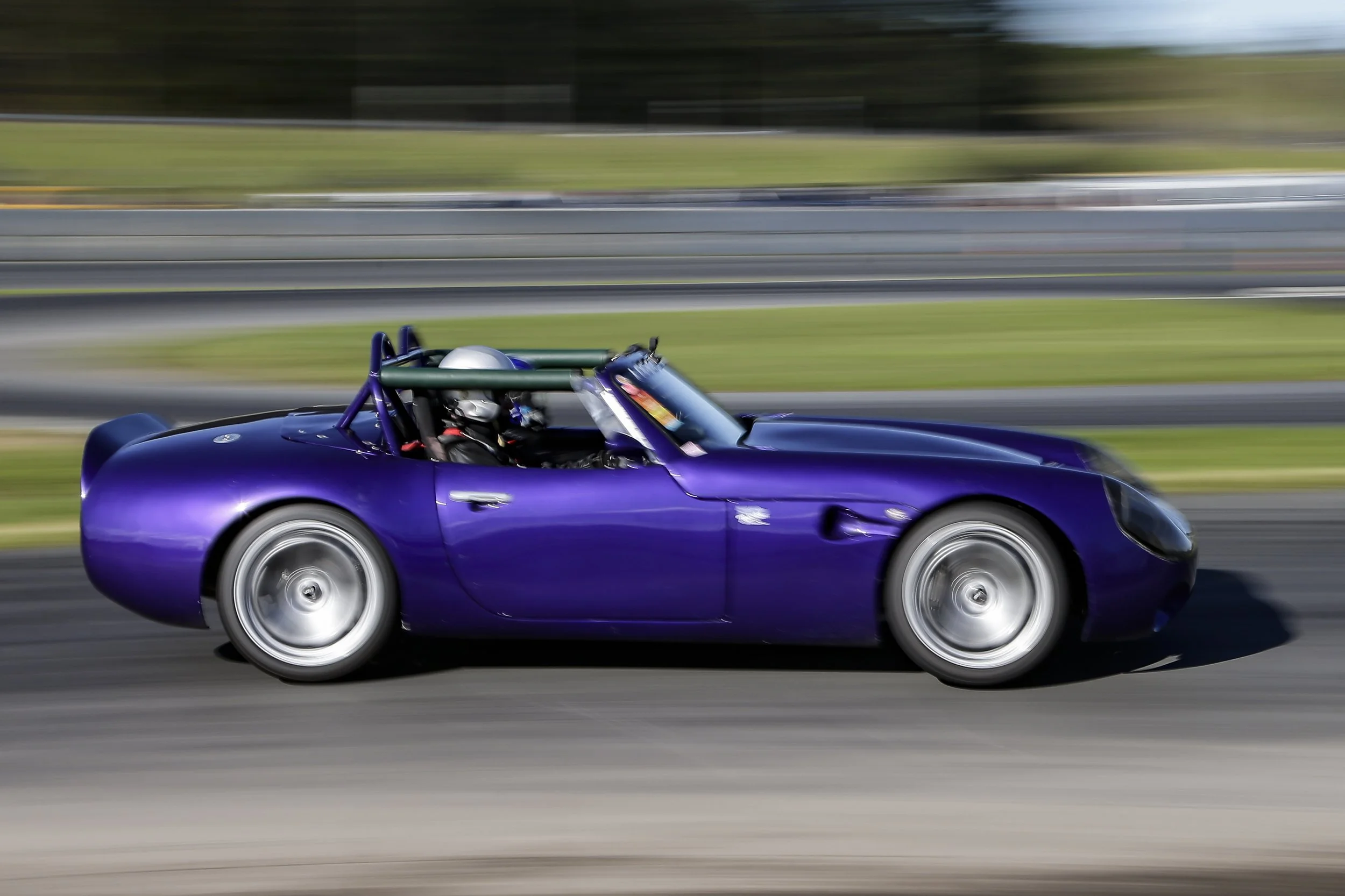 A purple sports car moving at high speed on a race track, with a driver wearing a helmet inside.