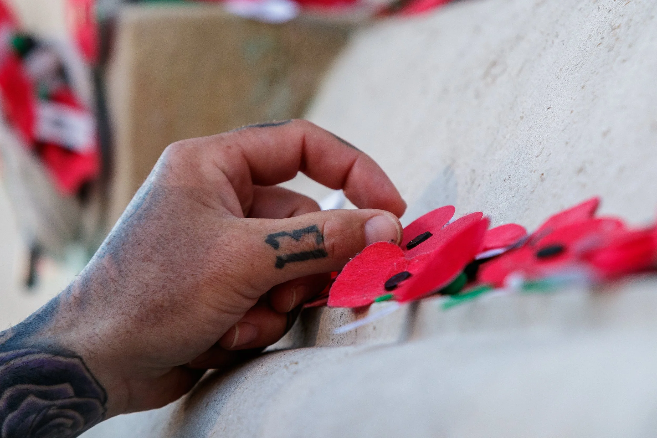 Close-up of a person's hand placing red and green paper poppies on a white surface, with some tattoos visible on the hand.