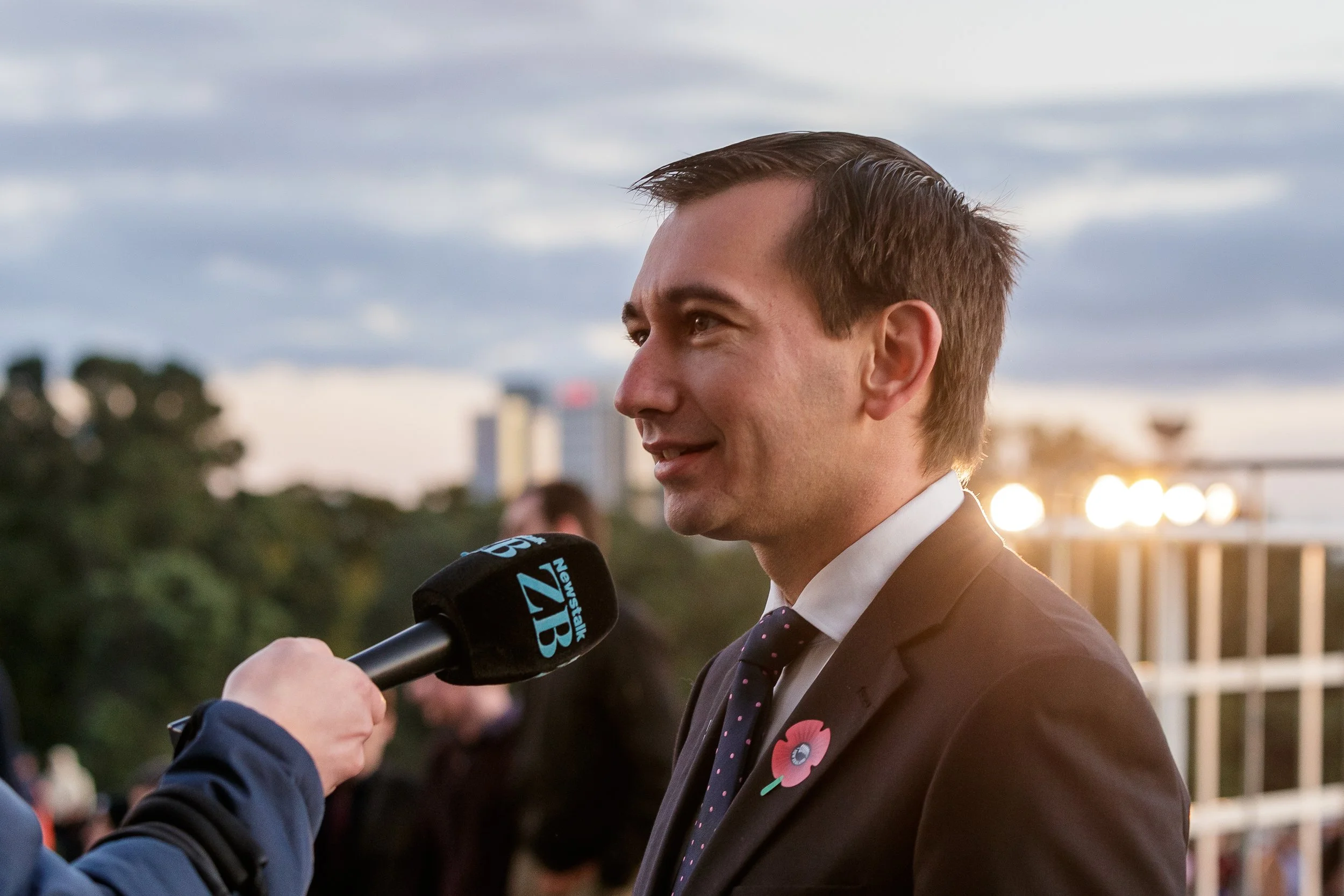 A man being interviewed with a microphone outdoors wearing a brown suit and a pink poppy pin, with a city skyline and cloudy sky in the background.