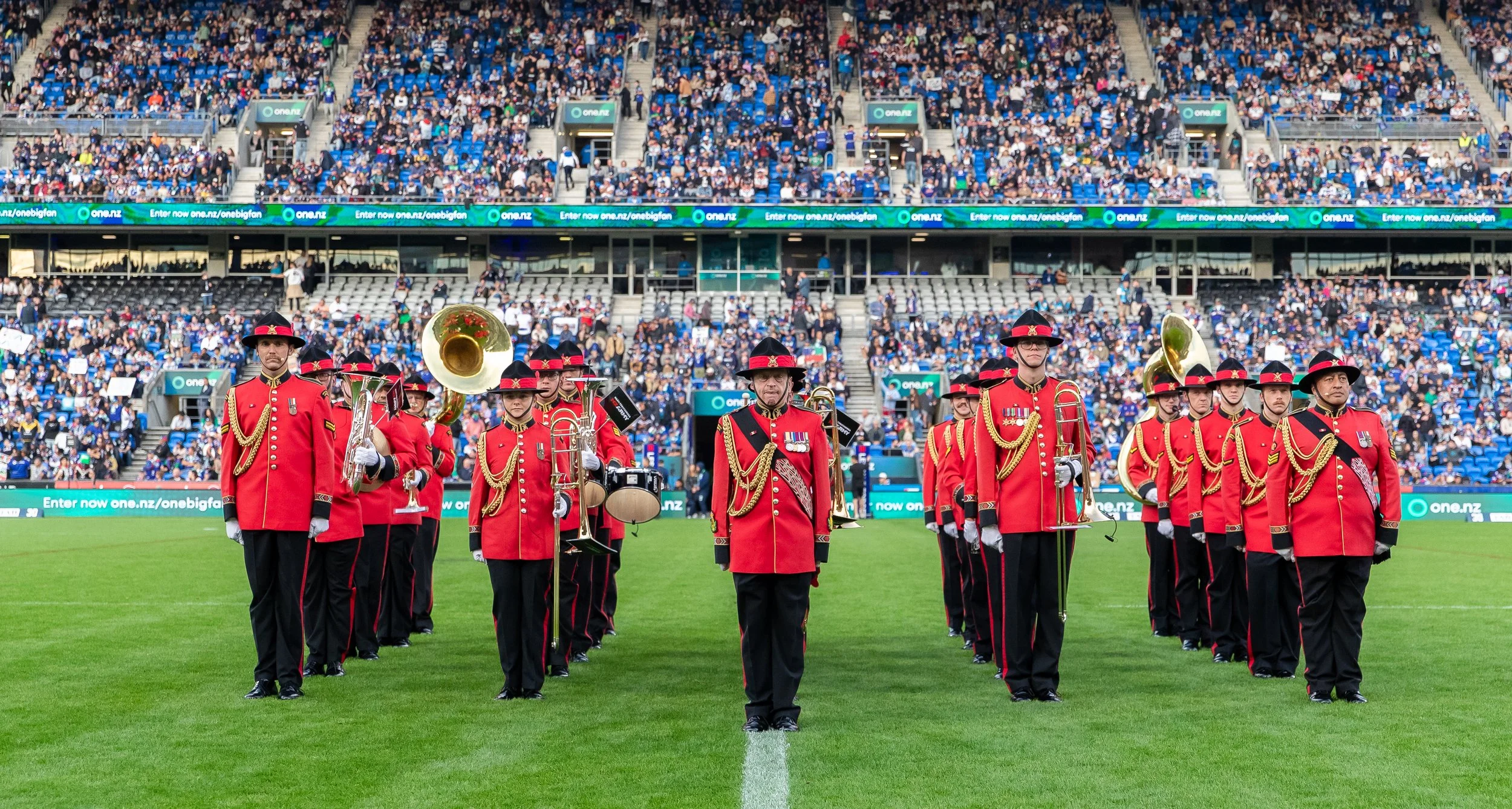 A marching band in red uniforms performing on a sports field with a large seated crowd in the stadium background.