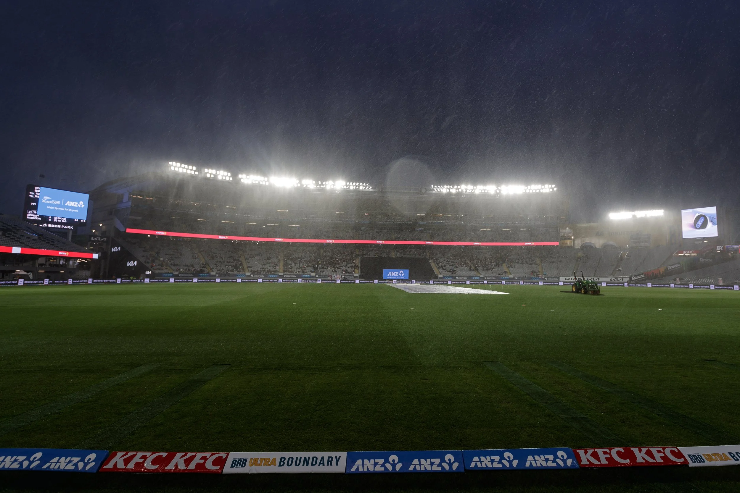 A large sports stadium under rain at night with bright floodlights, electronic scoreboards, and advertising boards, including brands like KFC and ANZ, with a wet grass field and a small piece of equipment on the field.