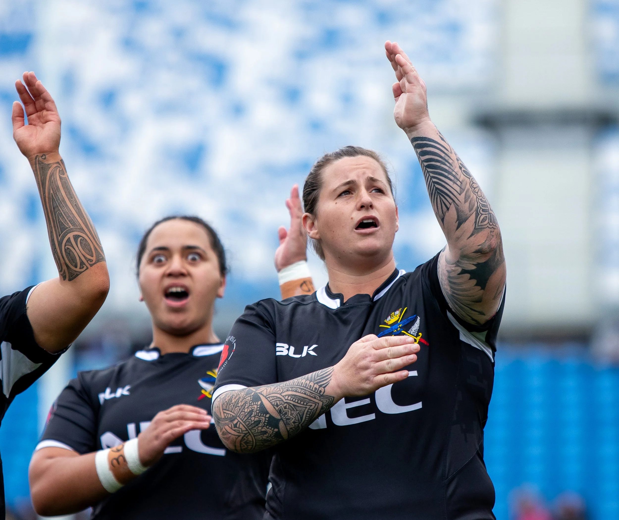 Line of rugby players standing with raised arms, wearing black jerseys, tattooed arms, and focused expressions, against a cloudy sky background.