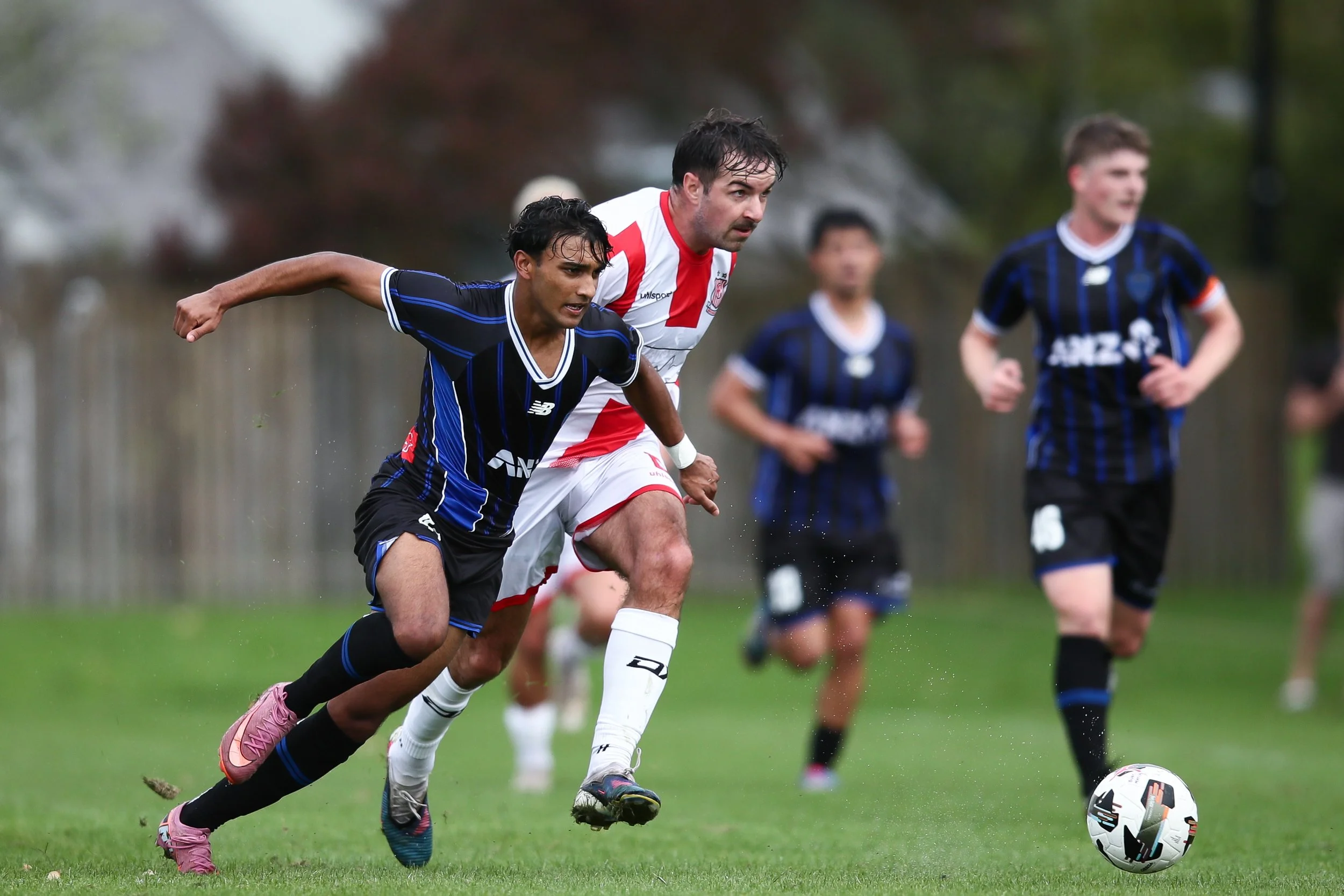 Two soccer players, one in a blue and black uniform and the other in a white and red uniform, chase after a soccer ball during a game on a grassy field, with additional players in the background.