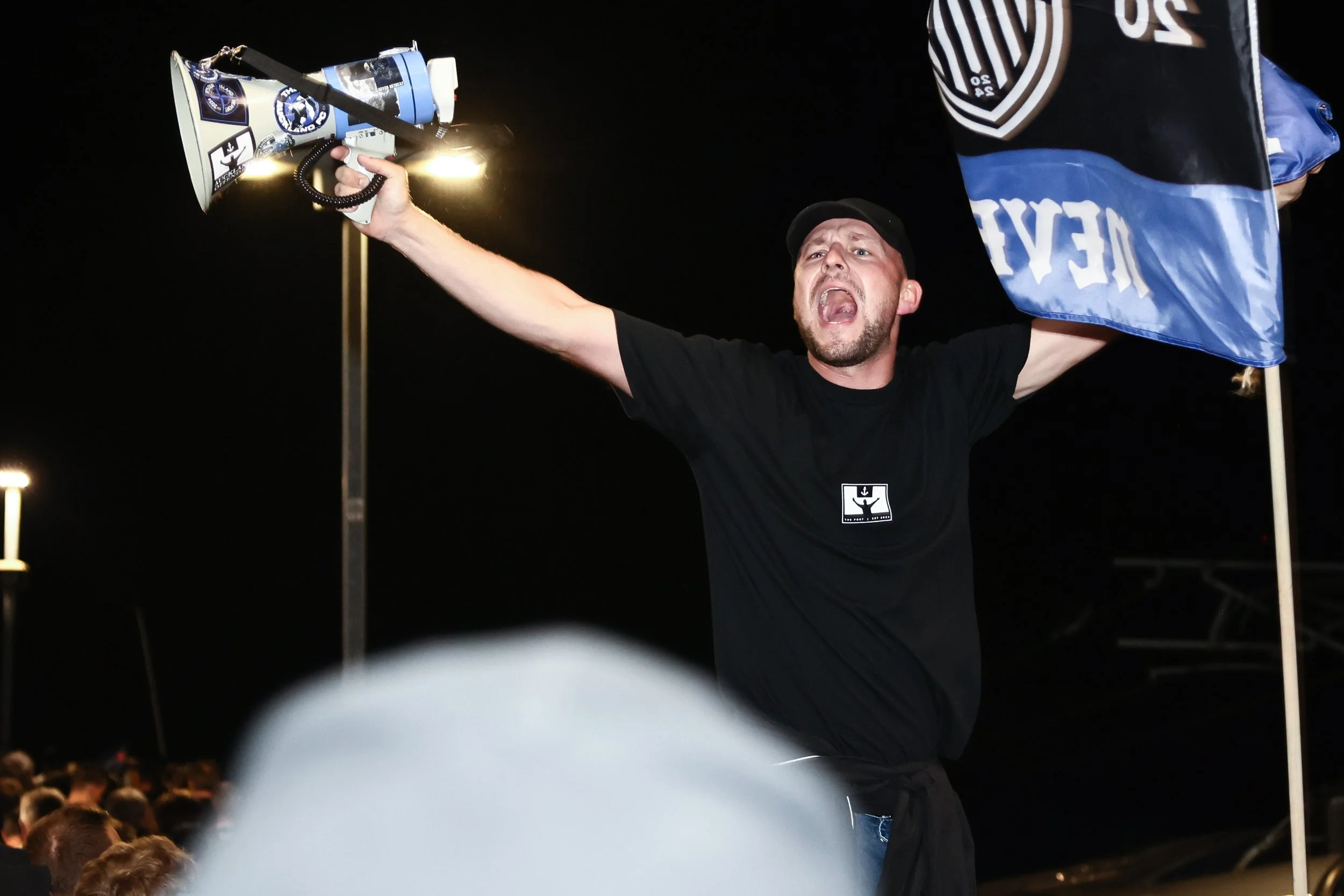 A man passionately protesting at night, holding a megaphone with sports-related stickers, and a flag. Crowd visible in the background.