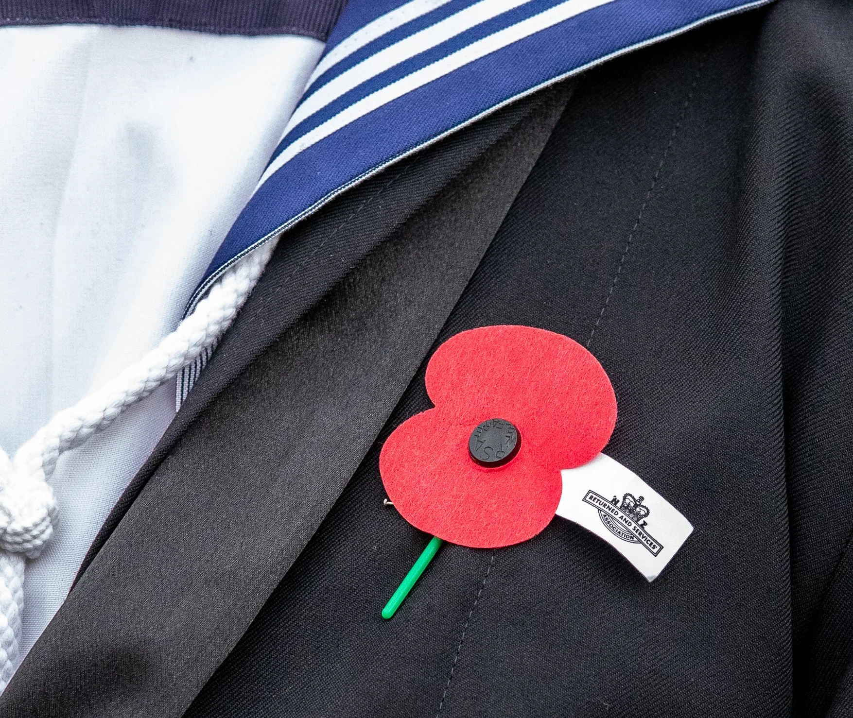 A close-up photo of a lapel with a red poppy flower attached, a black button, and a white clip with text, on a dark fabric background. The photo also shows part of a striped blue, white, and black tie and part of a white shirt liner.