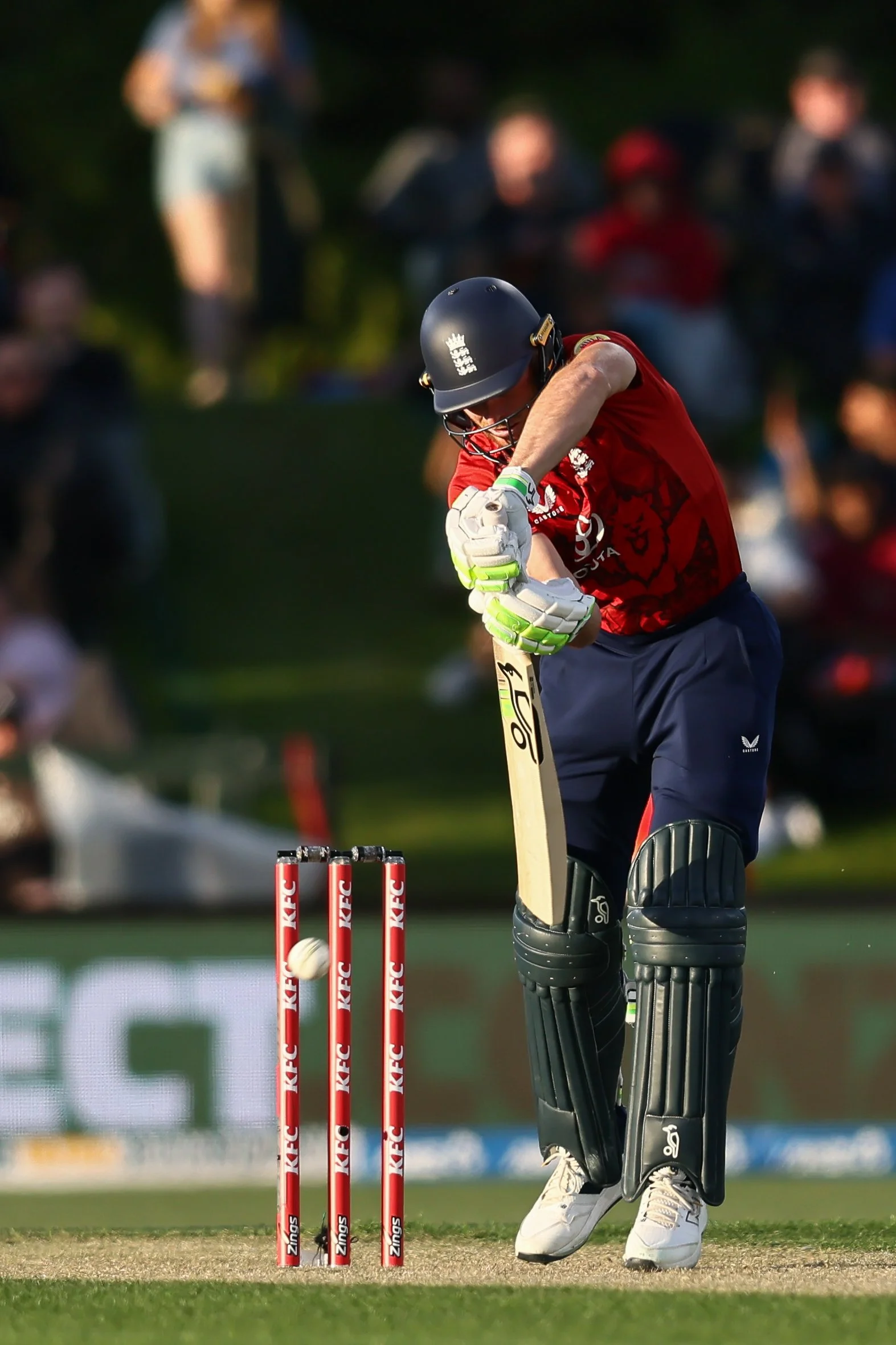 A cricket player in red and black uniform hitting a cricket ball during a match. The player is wearing protective gear, including a helmet, gloves, pads, and shoes. In the background, spectators are watching, and the wickets with red stumps are visib