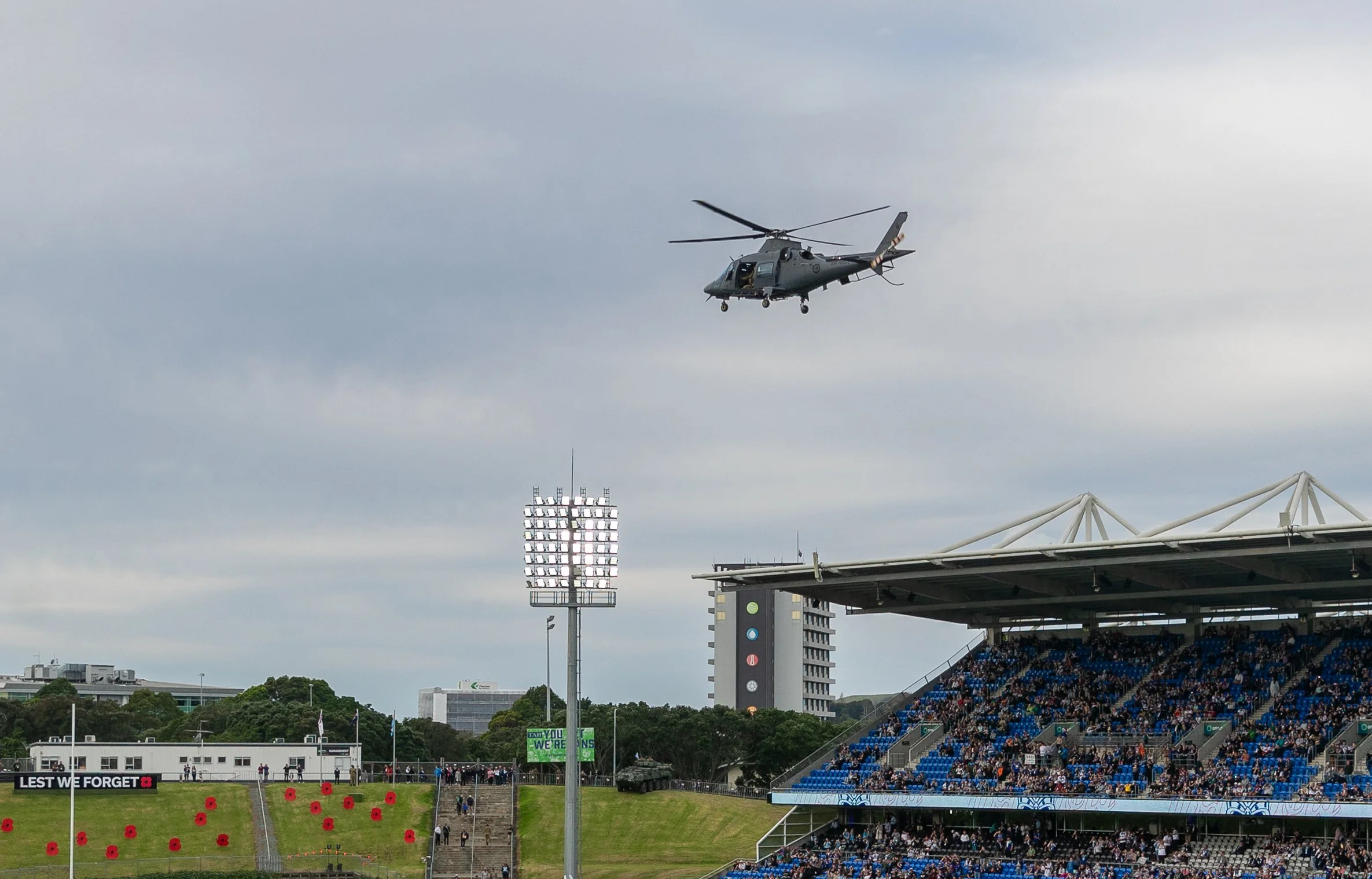 A helicopter flying over a sports stadium during a daytime event, with spectators in the stands and a large digital scoreboard visible.