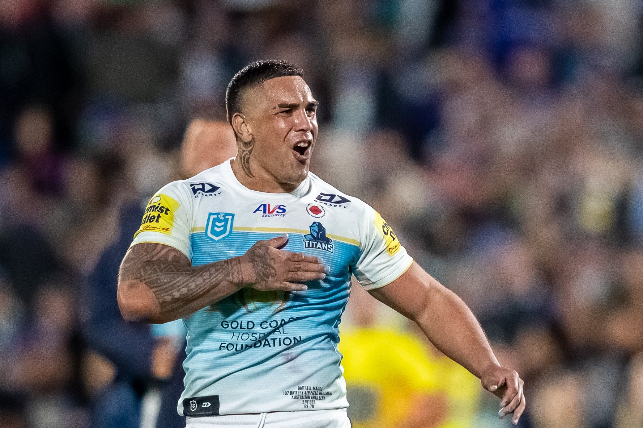 A rugby player wearing a white and light blue jersey with various sponsor logos, including Gold Coast Hospital Foundation, is in a moment of celebration or intense emotion during a game, with one hand on his chest and his mouth open.