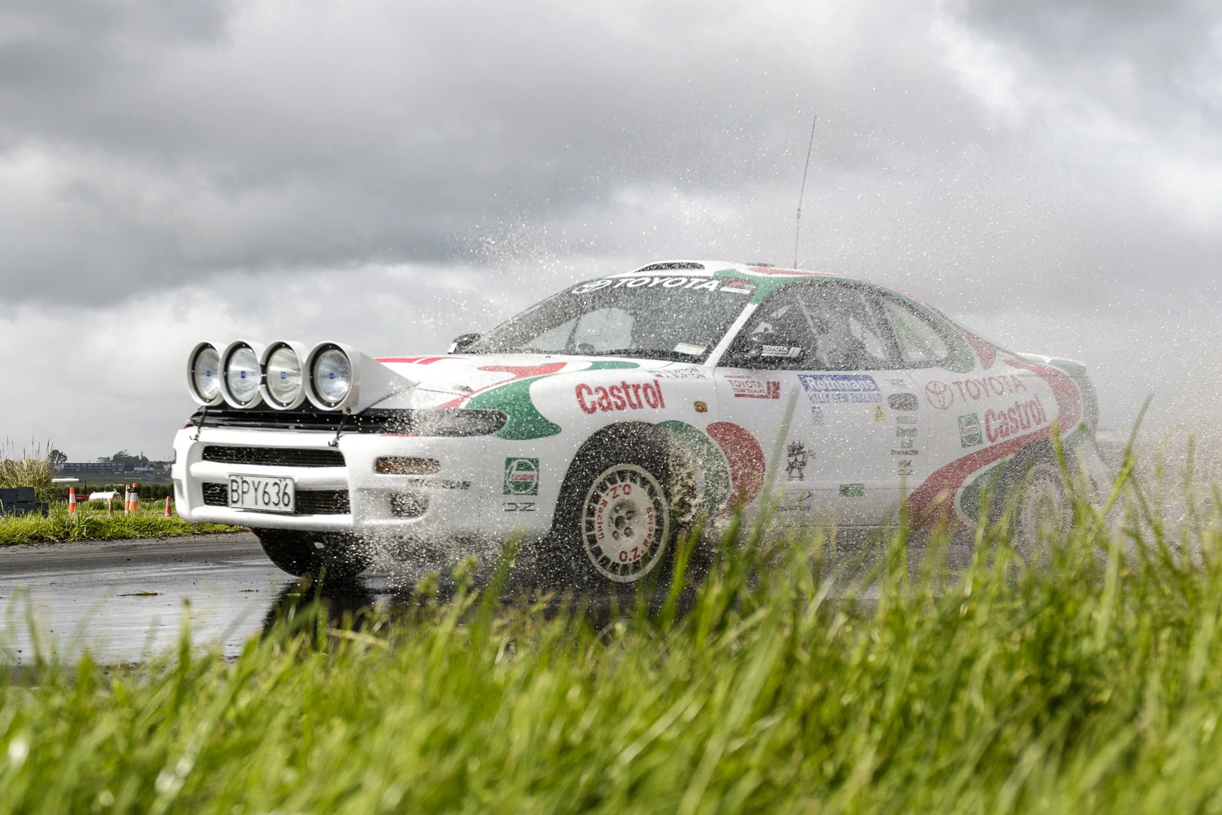 A race car speeding through a wet track with spray coming off its wheels, against an overcast sky.