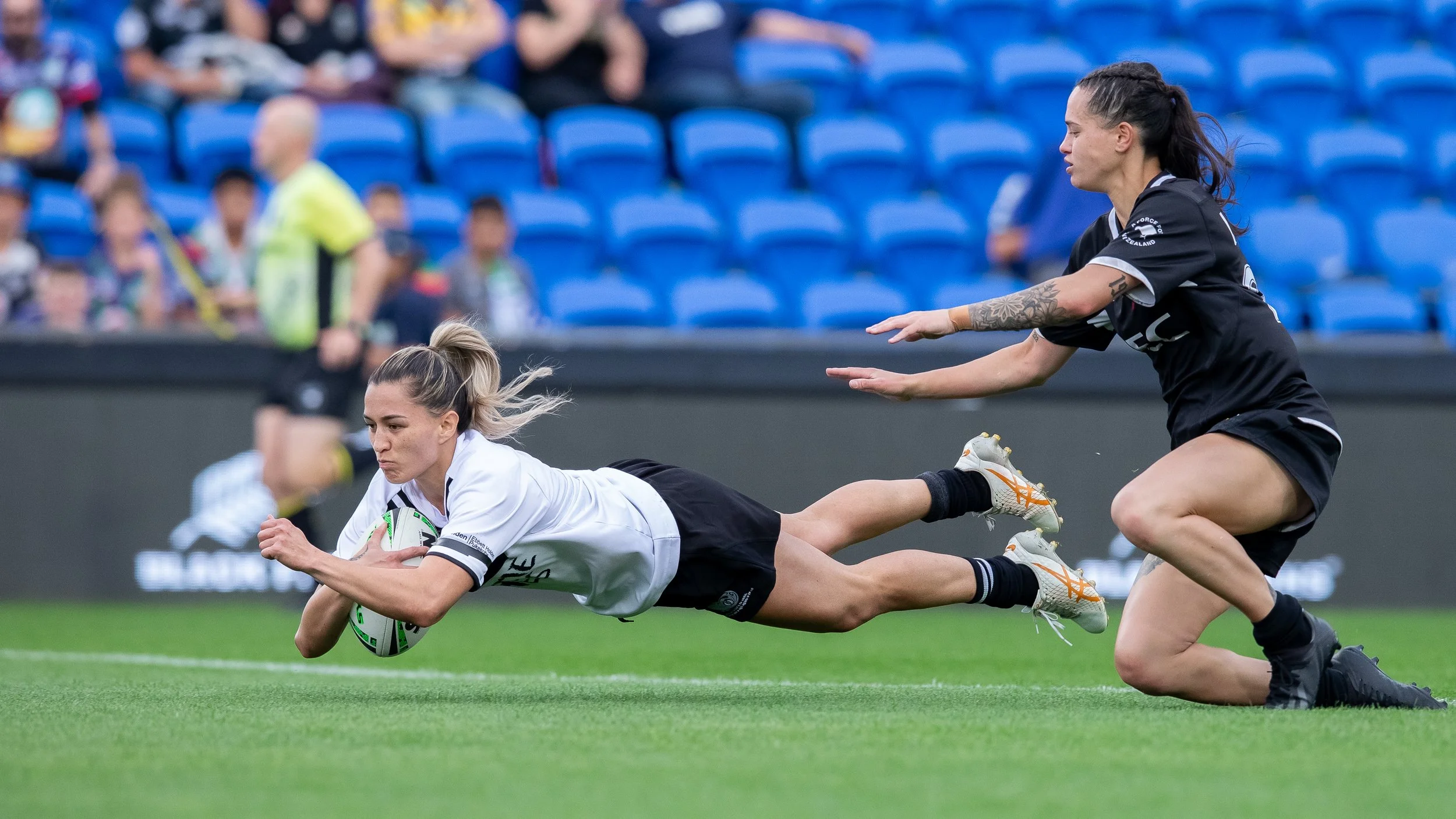 A rugby player in white uniform diving forward to catch a rugby ball, while a player in black uniform attempts to block or tag her during a match on a grass field.