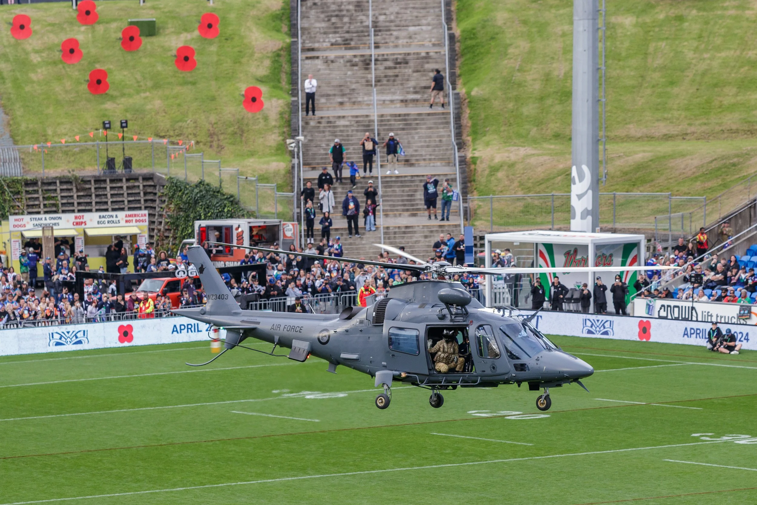 A military helicopter on the field with soldiers inside visible through the cockpit. In the background, there are crowds of people and a stadium seating area with stairs and a grassy slope decorated with red poppy symbols.