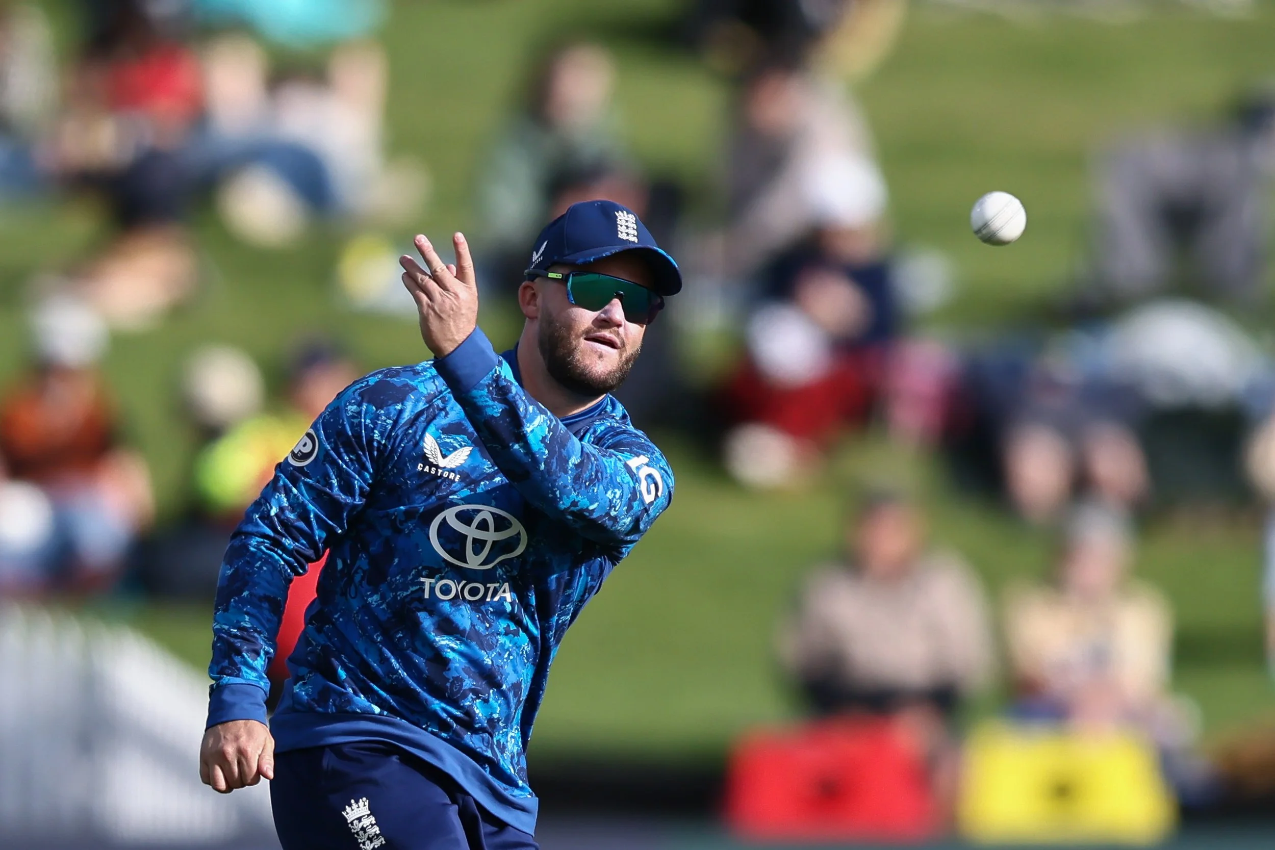 A male cricket player wearing sunglasses, a blue patterned shirt with sponsor logos, and a navy cap, is preparing to catch or hit a cricket ball during a game. The background shows a blurred crowd and green field.