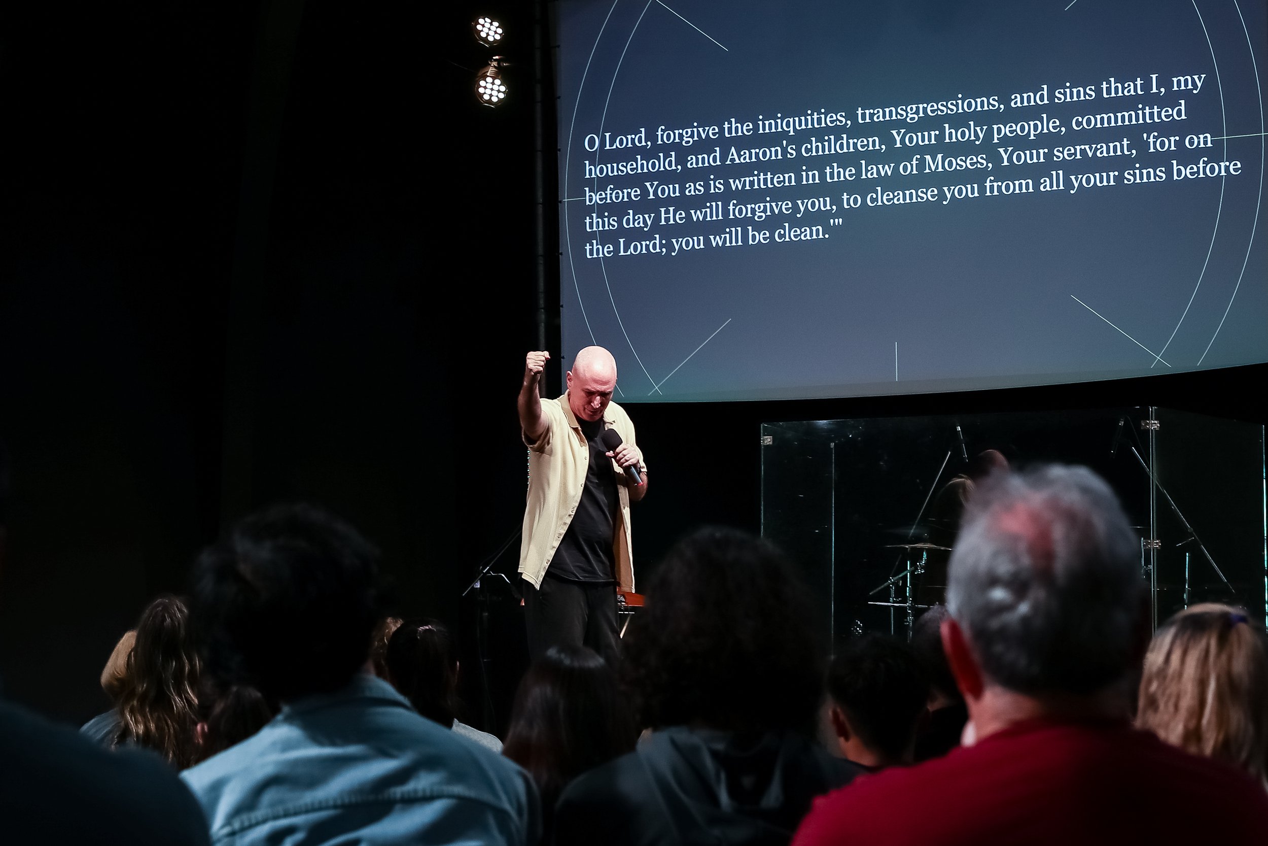 A speaker giving a presentation in front of a large screen displaying a prayer text, with an audience seated and listening attentively.