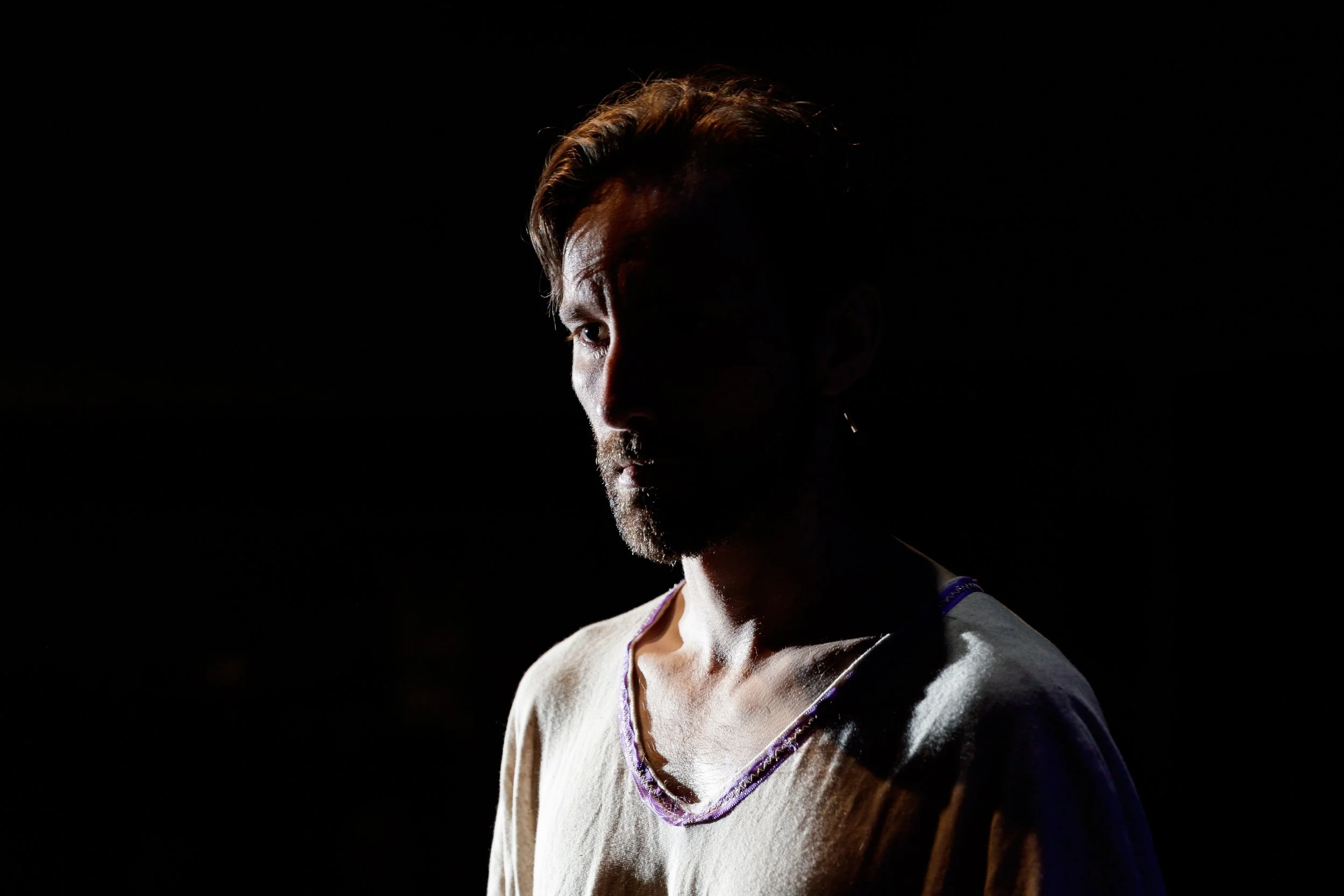 A person with short hair and a beard, wearing a light-colored shirt with purple accents, is illuminated on one side against a black background.