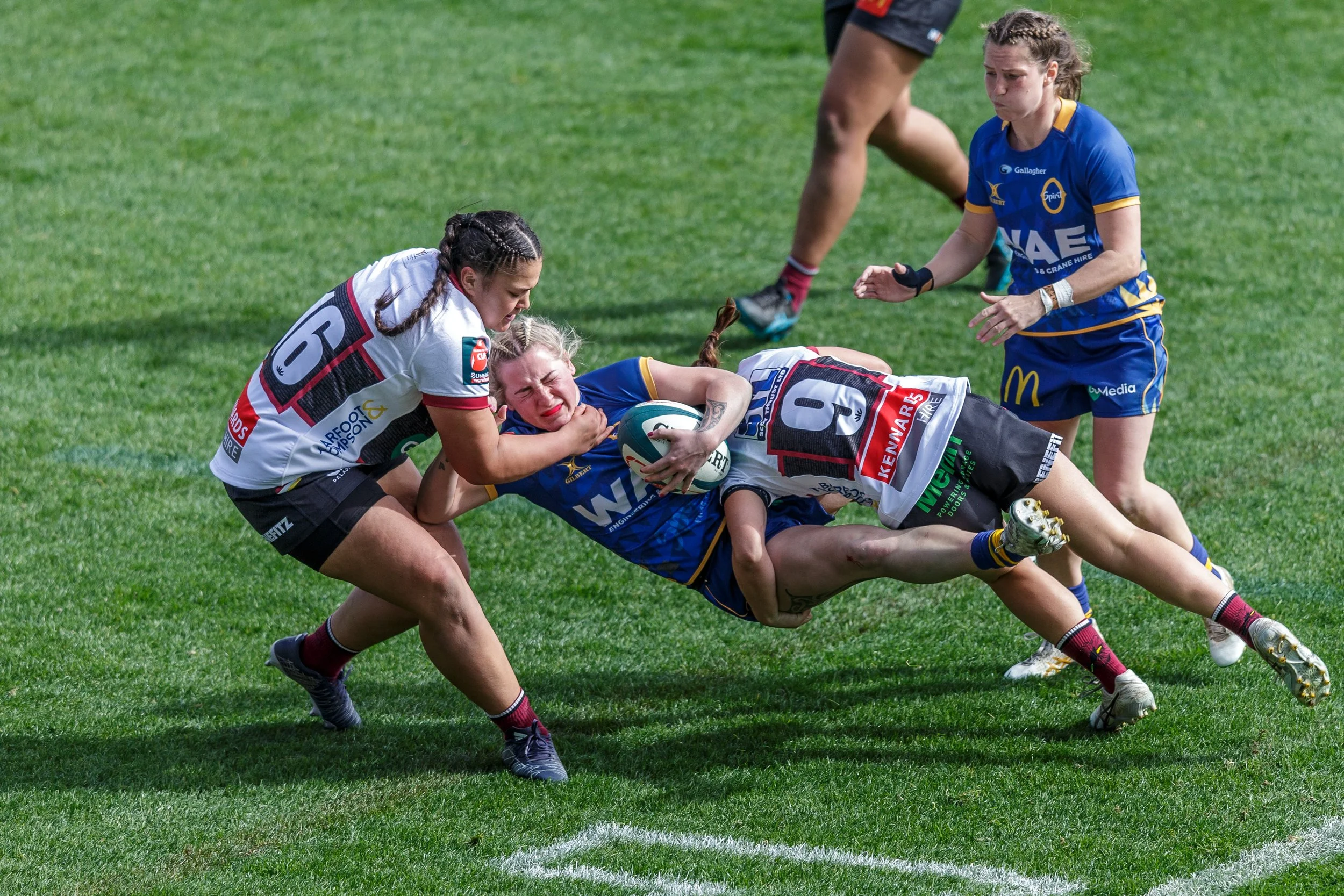 Women playing rugby on a grassy field, one woman in a blue jersey is tackled by two women in white and black jerseys, with another woman in a blue jersey nearby.