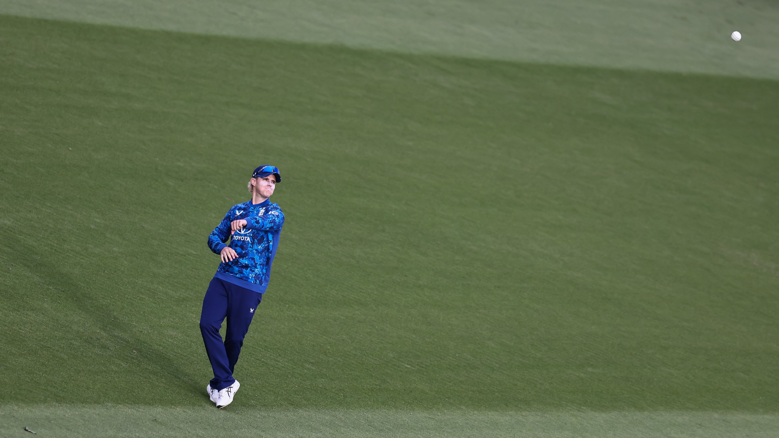 A person in a blue sports jacket and navy pants on a large grassy field, with a small white ball in the air nearby.