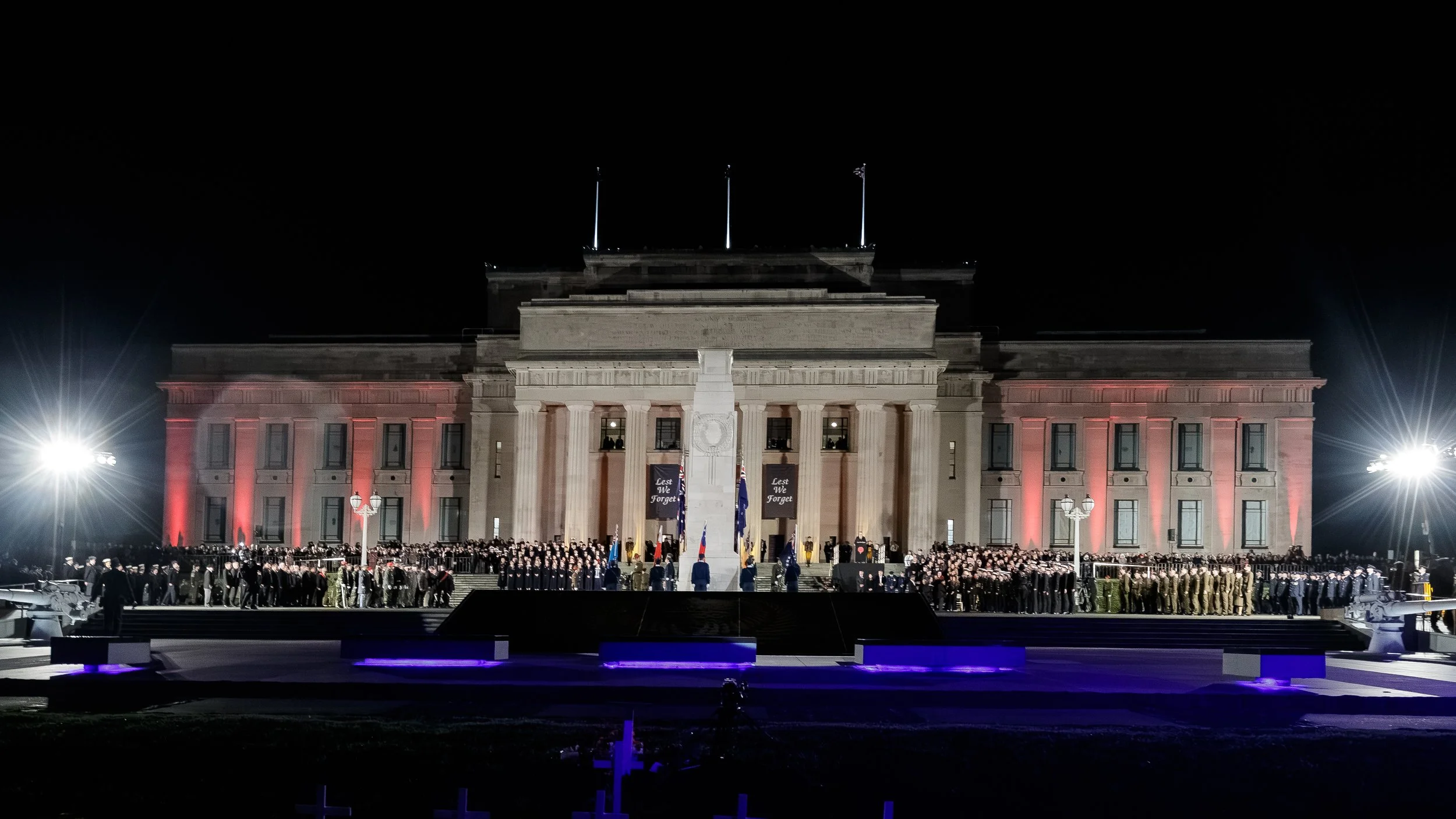 Nighttime military ceremony in front of a historic government building illuminated with red lights, with a large assembly of uniformed personnel and flags.