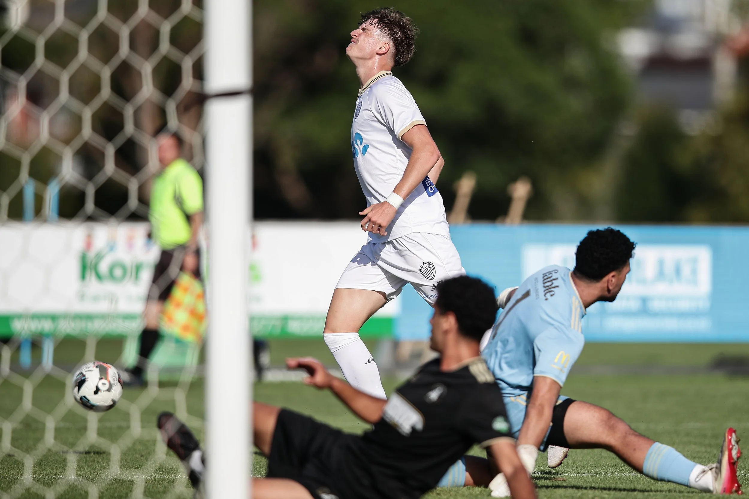 A soccer player in a white uniform is seen reacting near the goal post during a match, with an opponent in a light blue jersey lying on the ground nearby.