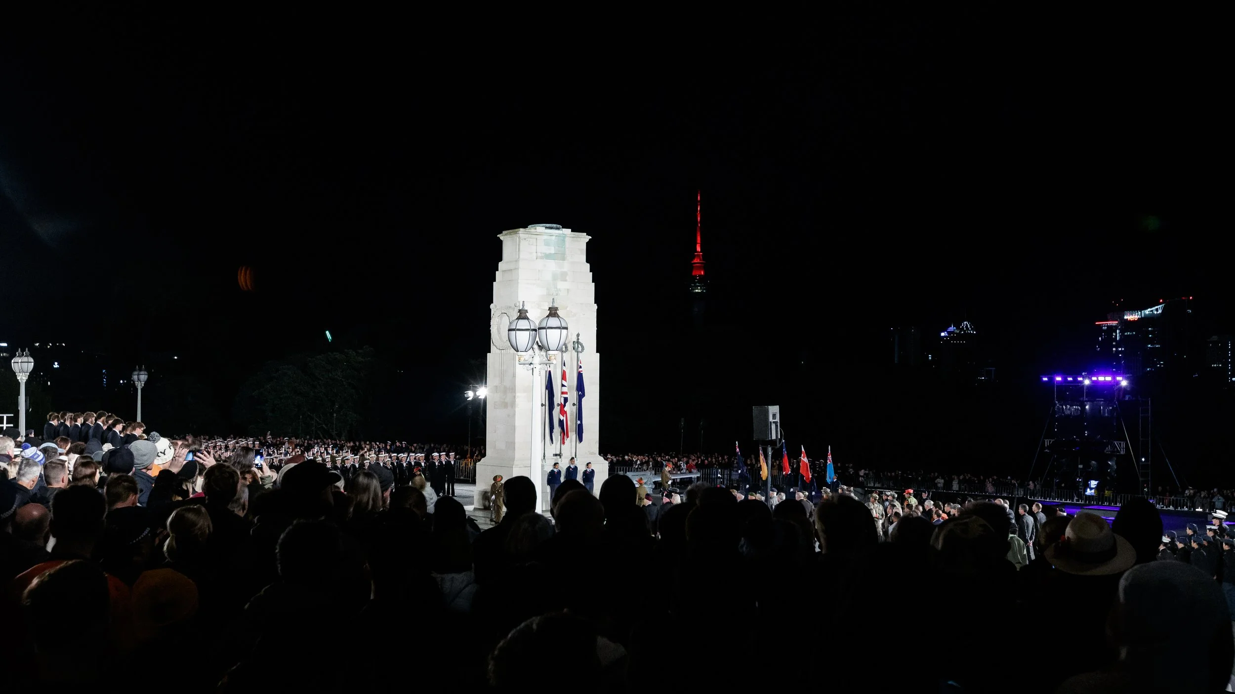 A night scene with a large crowd gathered at a memorial or monument, with flags and uniformed personnel in front of a white stone structure, illuminated by bright lights. A tall, illuminated TV tower is visible in the distance against the dark sky.
