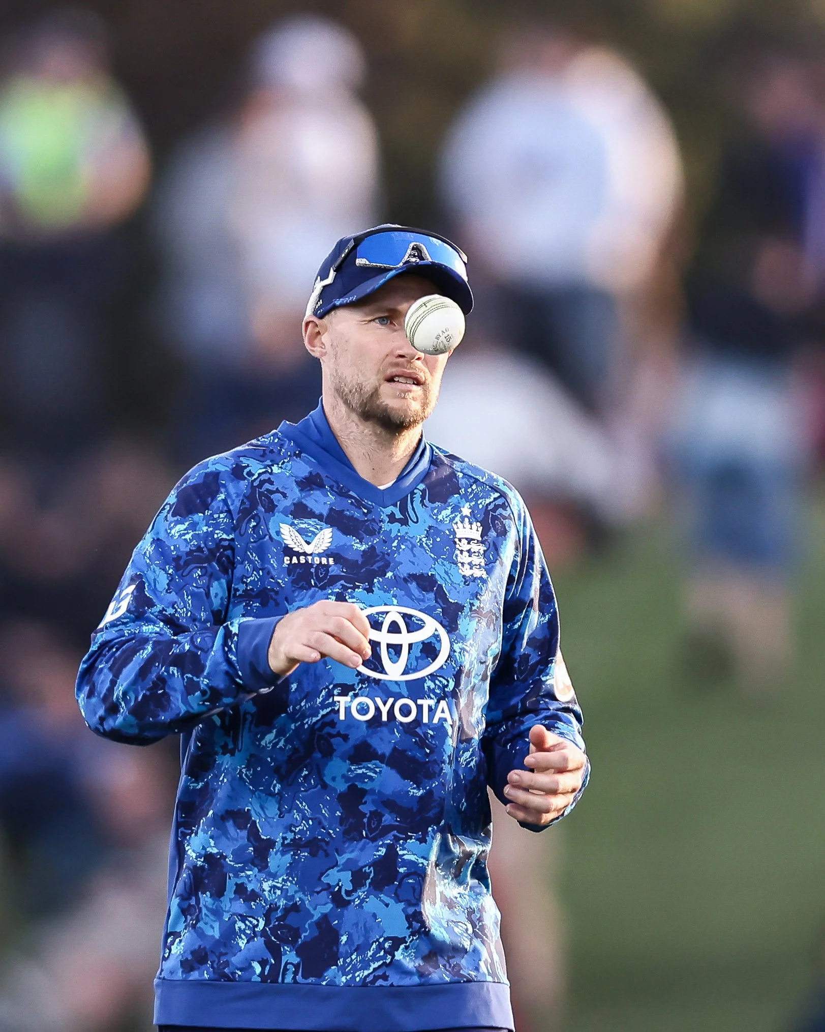 A male athlete wearing a blue sports jersey is on a cricket field. He is tossing a cricket ball in the air. The background shows out-of-focus spectators.