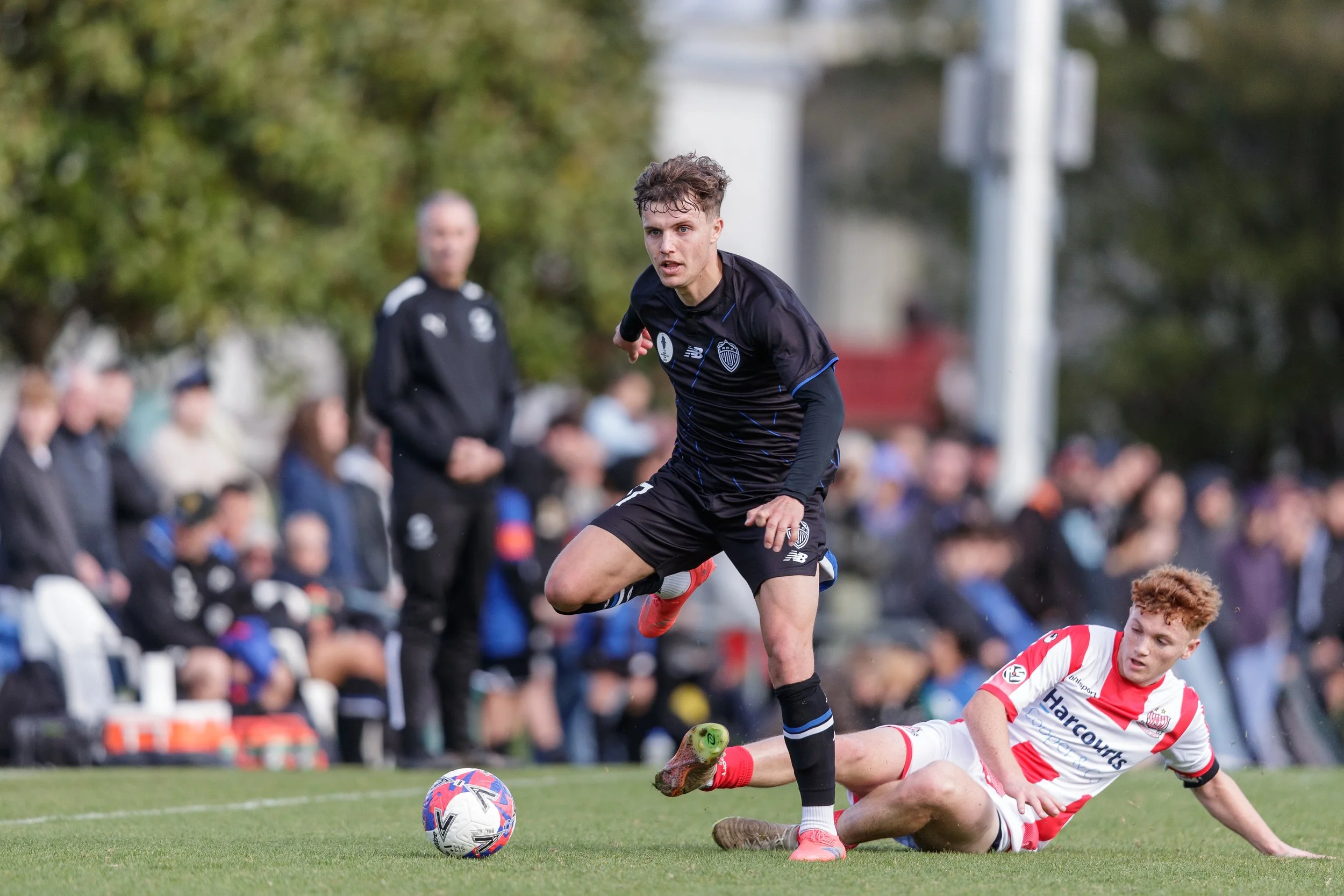 A soccer player in black uniform attempting to kick a soccer ball while another player in white and red uniform falls on the ground during a match with spectators in the background.