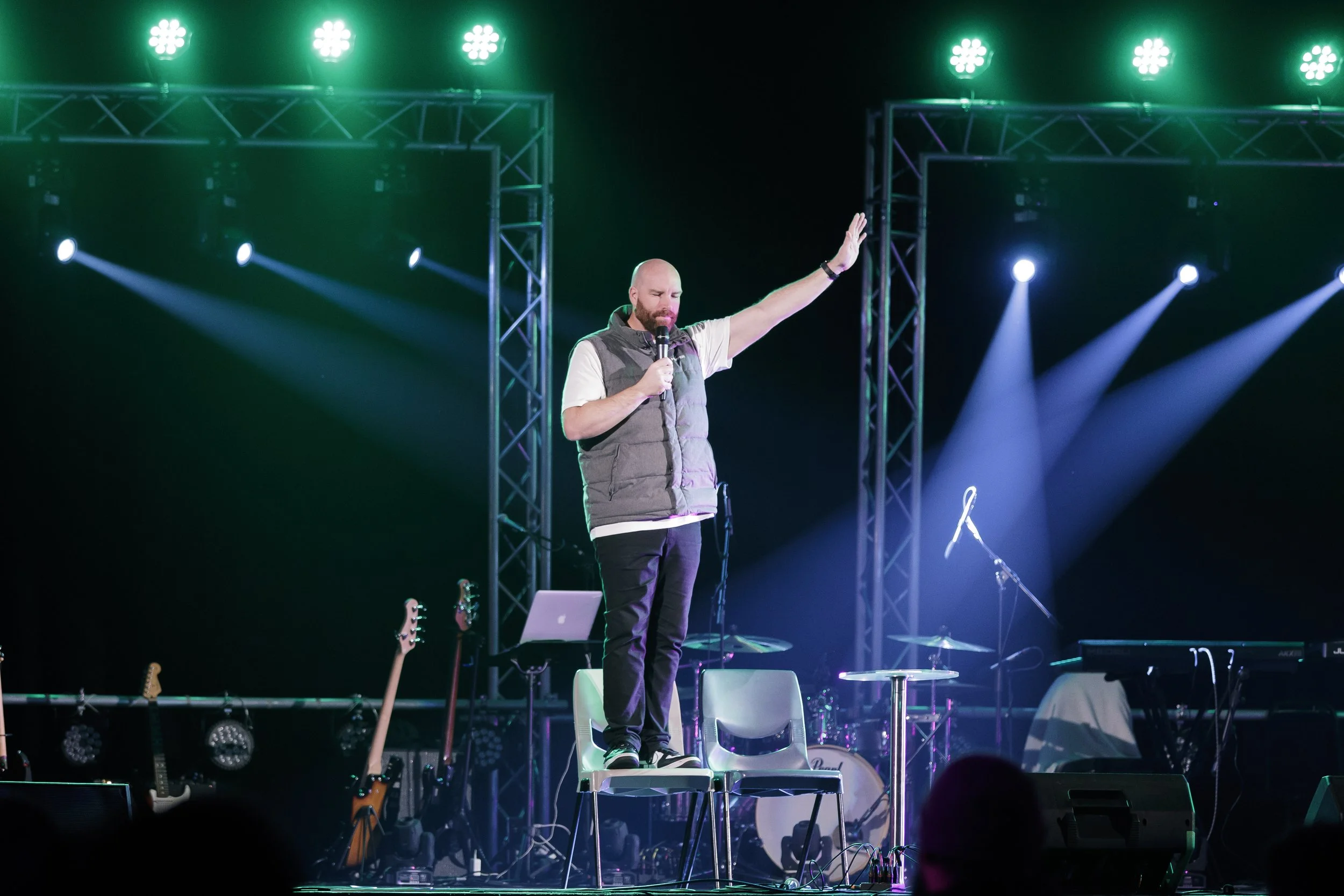 A man with a beard and a bald head is standing on stage holding a microphone and raising his right hand. The stage is lit with green and blue lights, with several guitars lined up in the background.