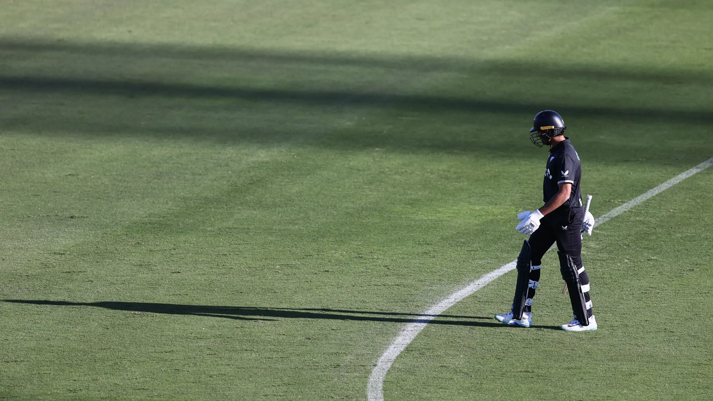 Cricketer standing on a cricket field, dressed in black sportswear, wearing a helmet and holding cricket gloves, looking contemplative.