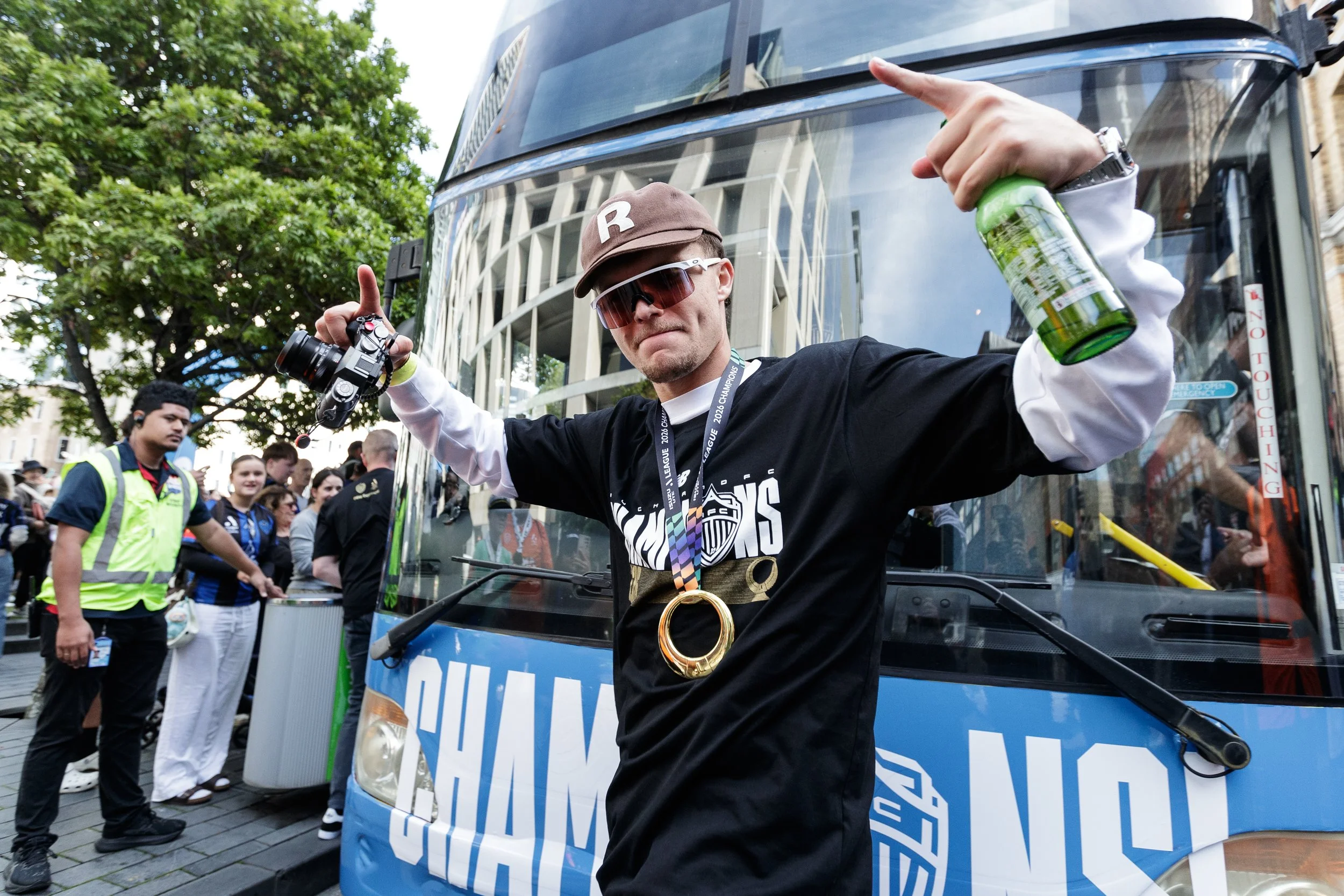 A man with sunglasses and a baseball cap standing in front of a bus, holding a water bottle and giving a thumbs-up, surrounded by a crowd at an outdoor event.