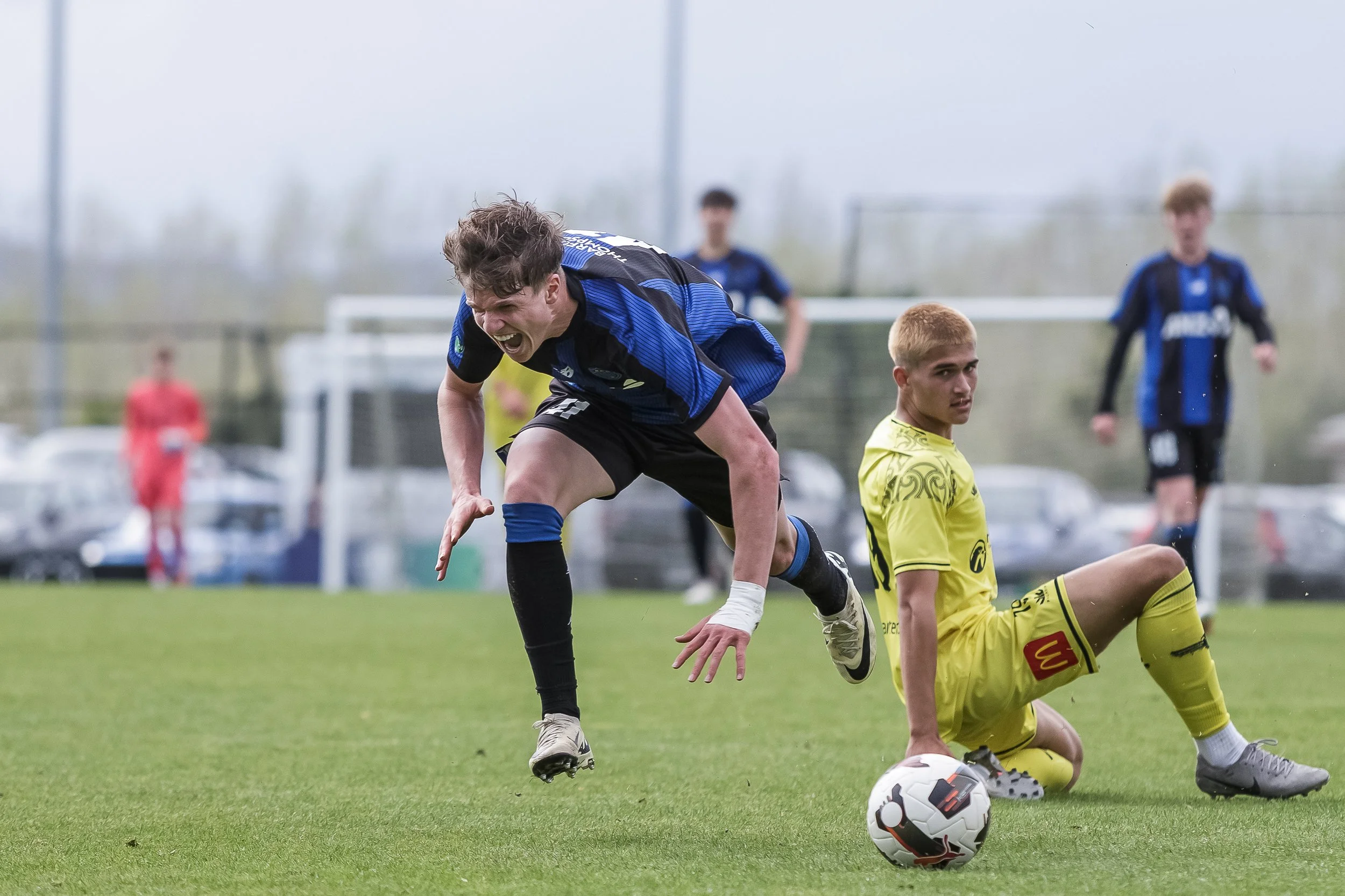 A soccer match with a player in a blue and black jersey, while a player in a yellow jersey sits on the field. Other players and a referee are visible in the background.