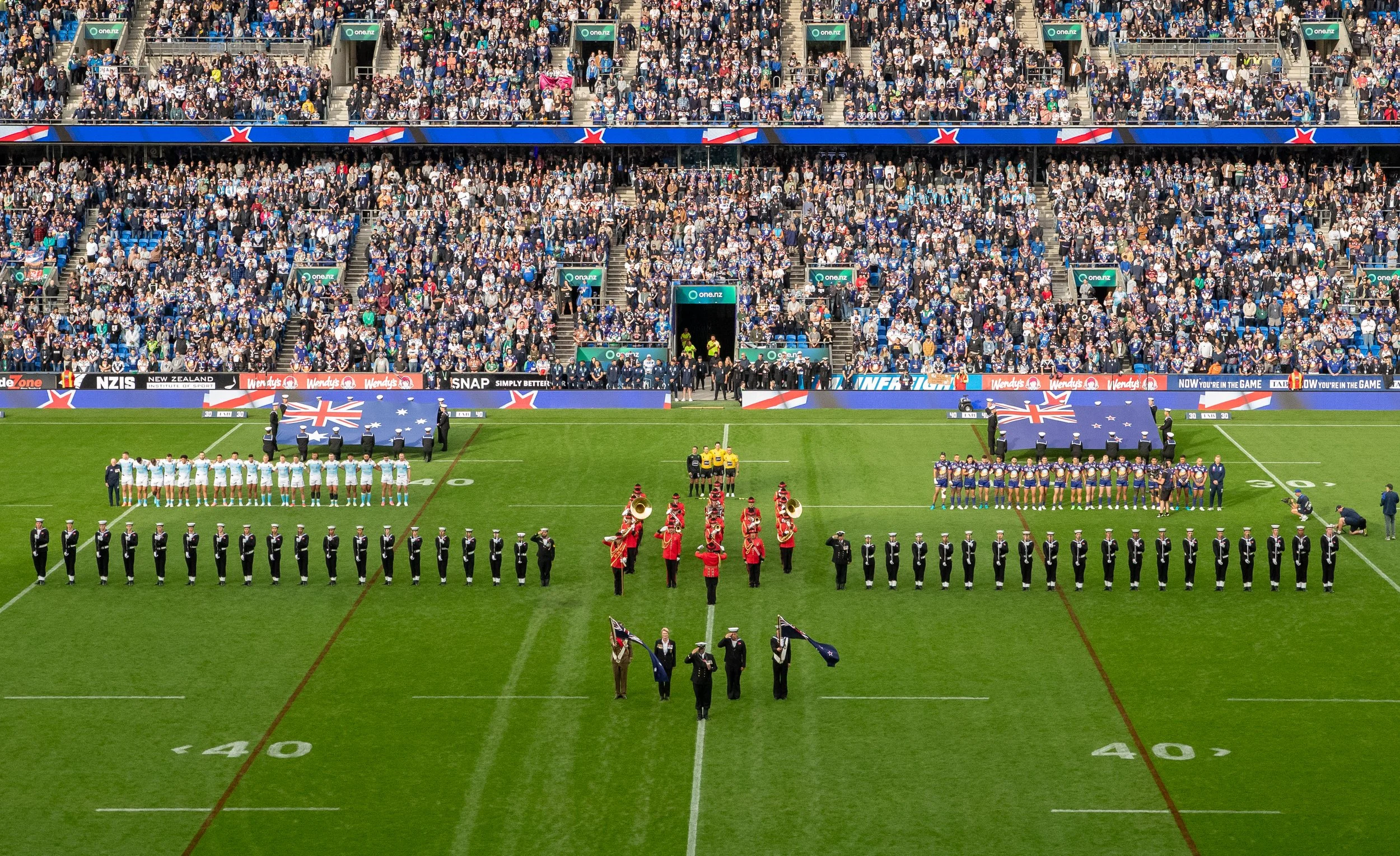 A packed stadium with an audience, flags, and players organized on the field for a pre-game ceremony at a rugby match, featuring two teams and uniformed personnel.